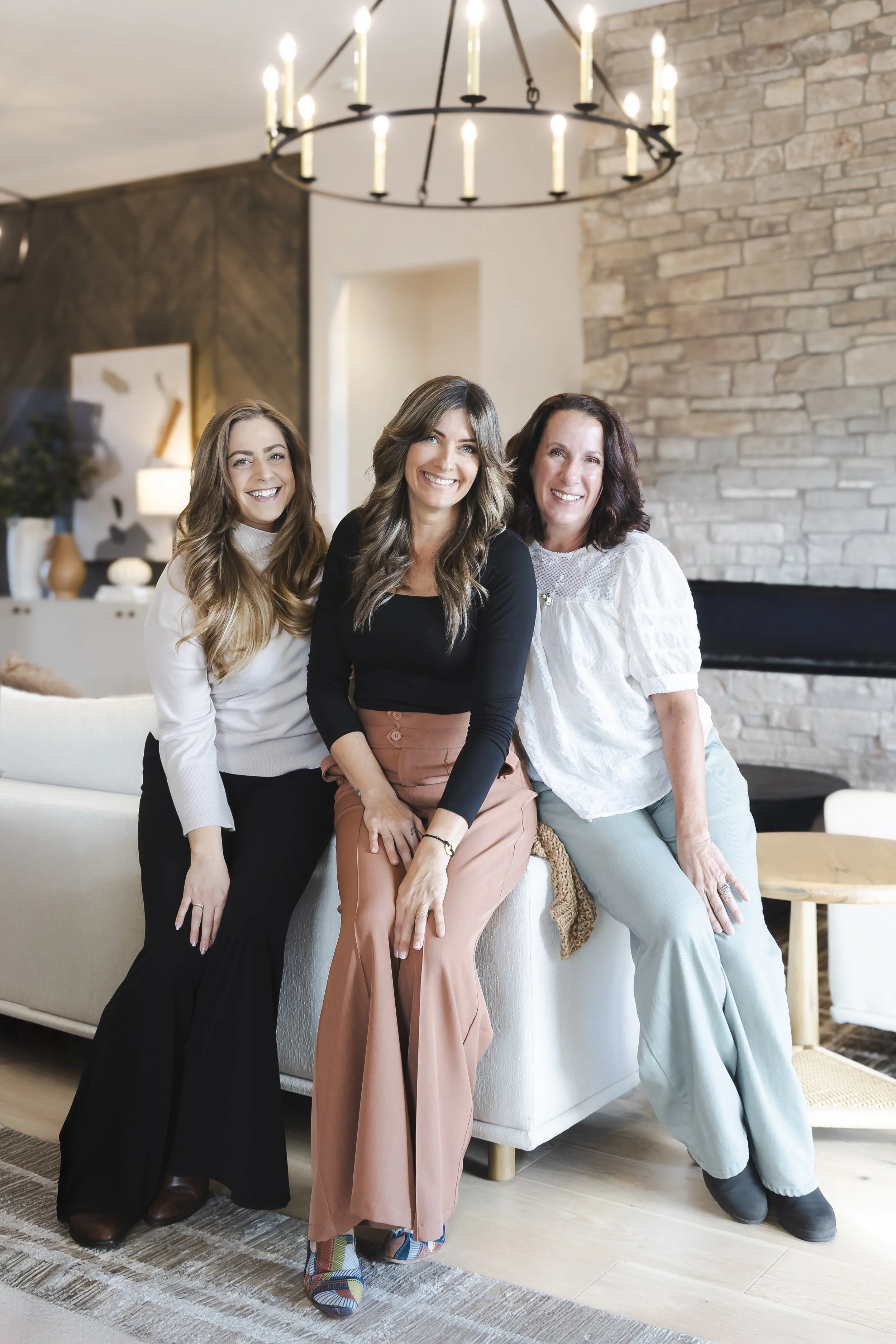 Three women smiling and sitting on a white couch in a cozy, modern living room.