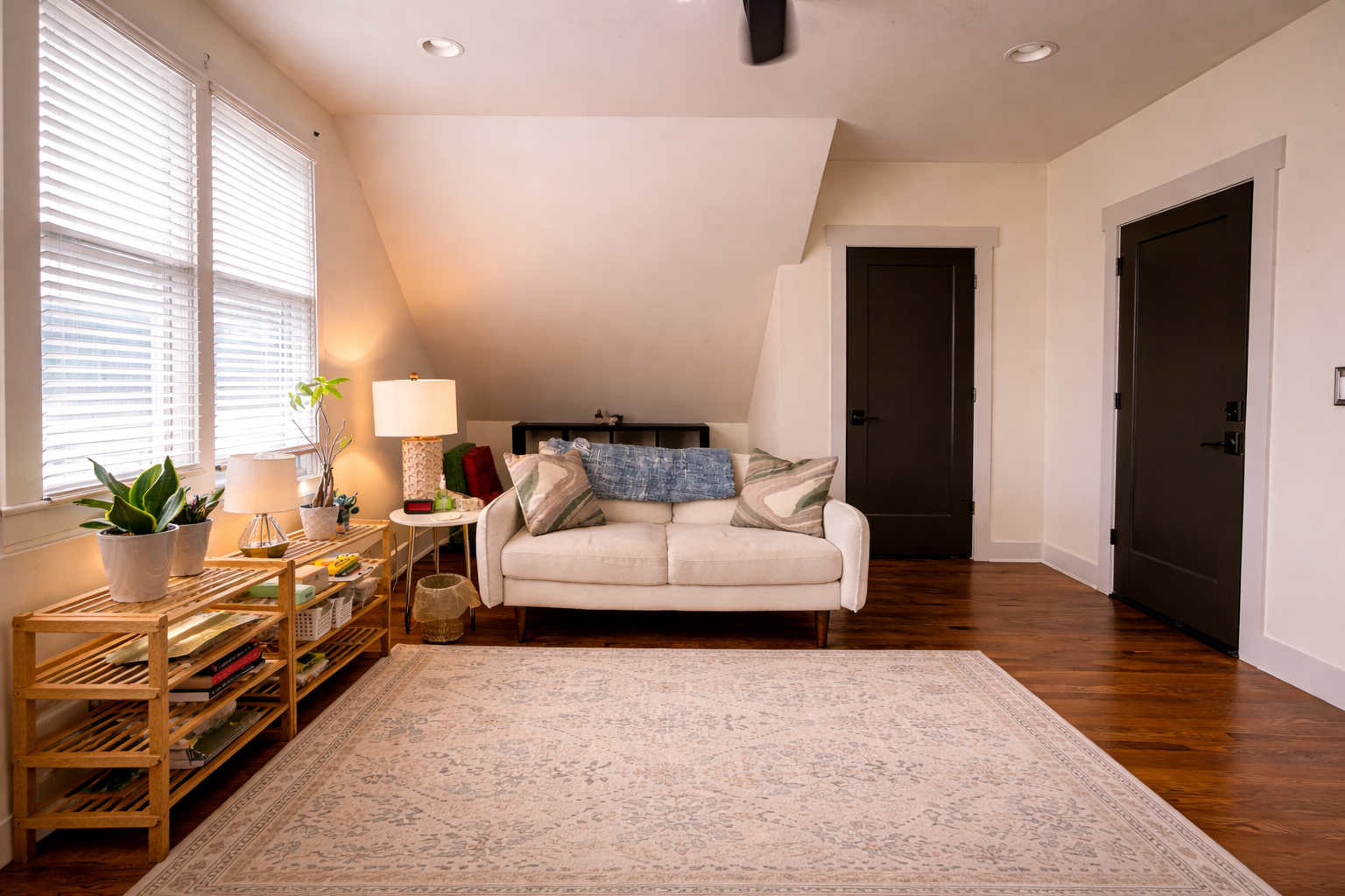 Living room with white loveseat, pillows, side table with lamp, wooden shelf with plants and books, large window with blinds, hardwood floors, and black doors.