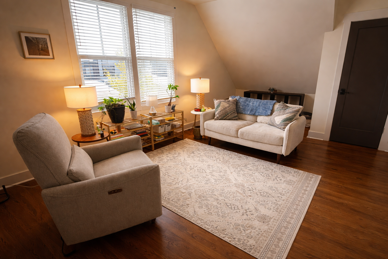 Living room with beige armchair and white sofa, wooden floor, patterned area rug, two side tables with lamps, window with blinds, and various decorative items and potted plants.