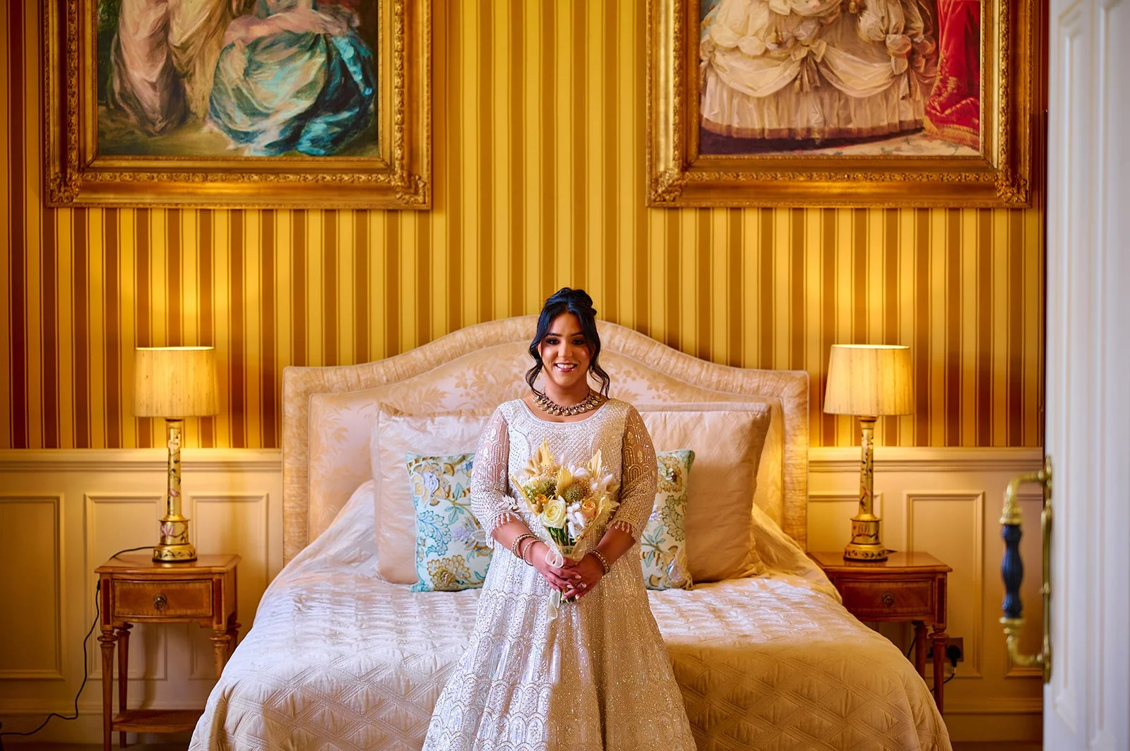 A bride cream-colored dress, holding a bouquet of flowers, standing in a luxurious bedroom with striped yellow wallpaper, large framed paintings, and matching lamps on bedside tables.