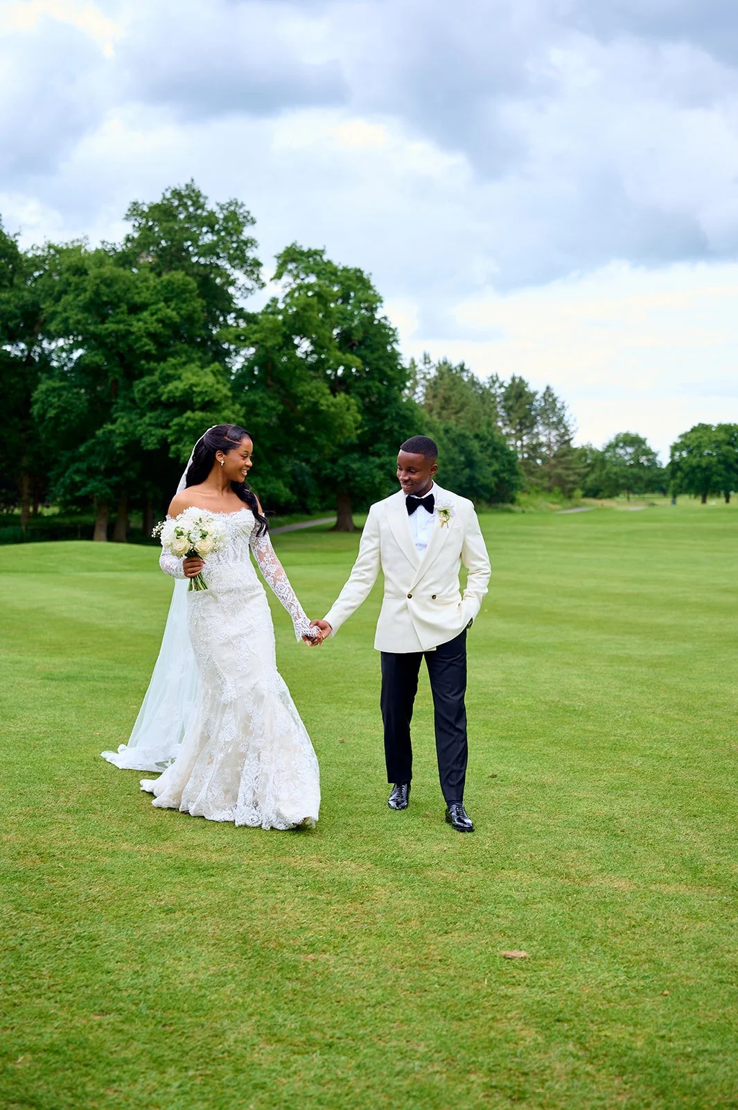 A bride and groom walking hand in hand on a grassy field, with trees and a cloudy sky in the background during a wedding photo shoot.