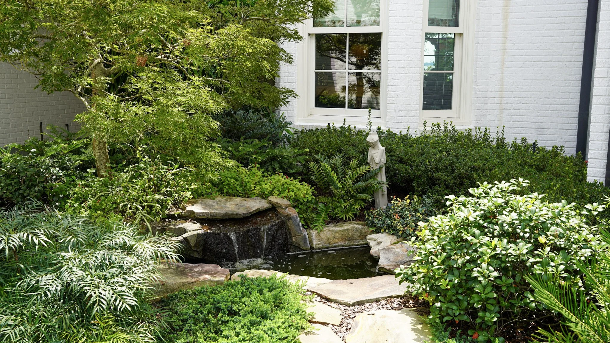 A well-maintained garden with a small pond, lush green plants, bushes, and trees in front of a white brick house with two large windows.
