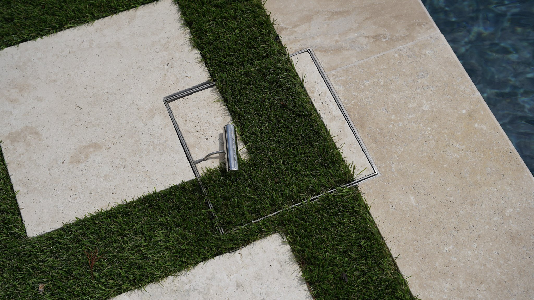 Close-up of a poolside drain cover surrounded by grass and beige stone tiles.