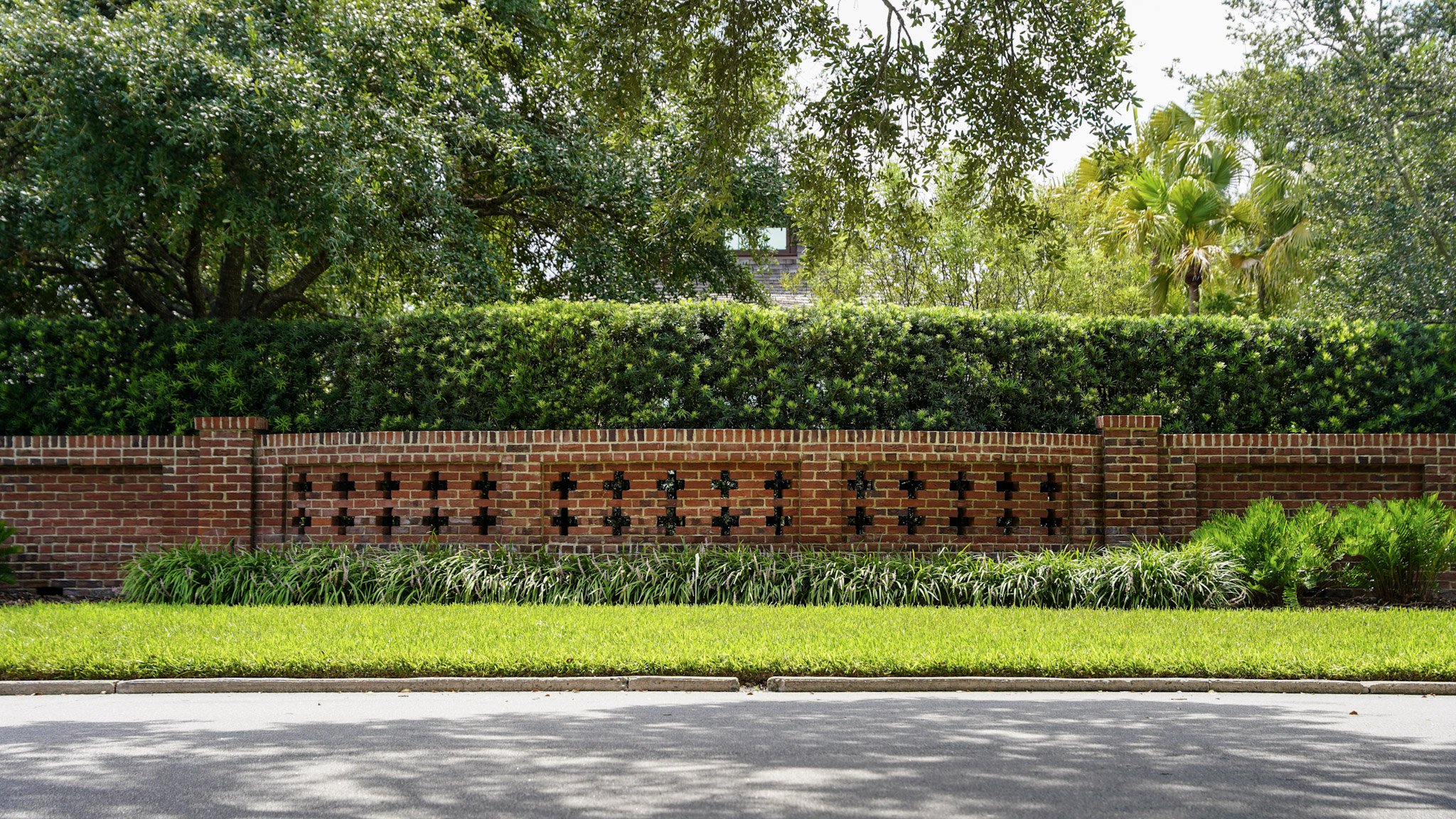 A brick wall with decorative openings, topped with trimmed greenery, and various trees and plants behind it, along a sidewalk and street.