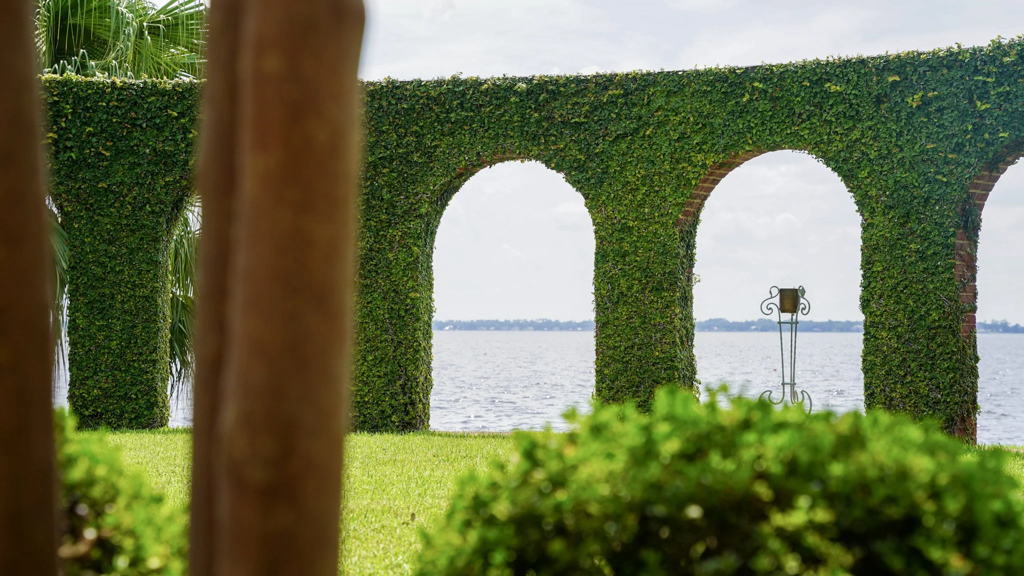 View of two large, arch-shaped structures covered with green ivy, overlooking a body of water in the distance, with a manicured lawn and a decorative metal stand in the foreground.