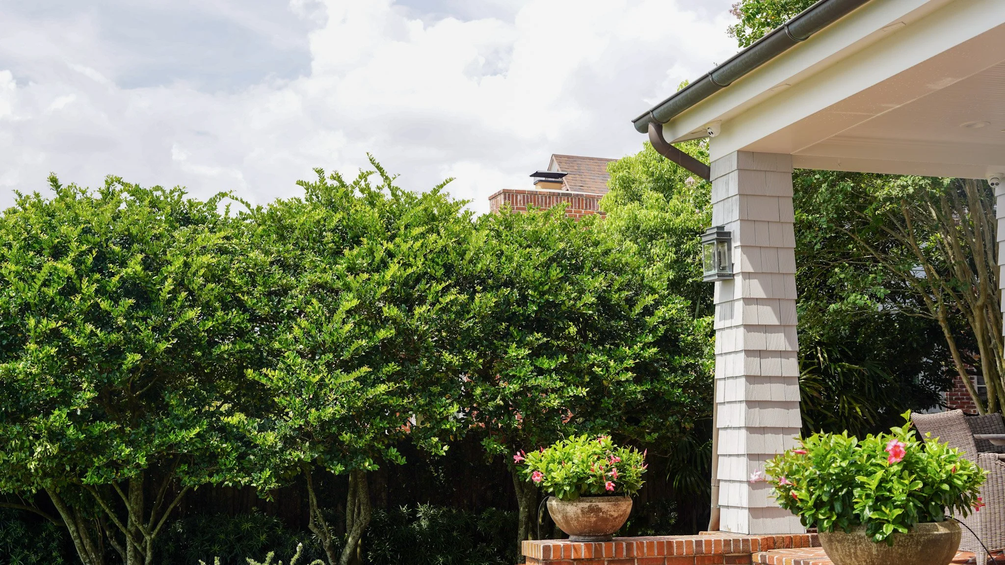 Side view of a house patio with tall green bushes, potted pink flowering plants, and a white porch column, under a cloudy sky.