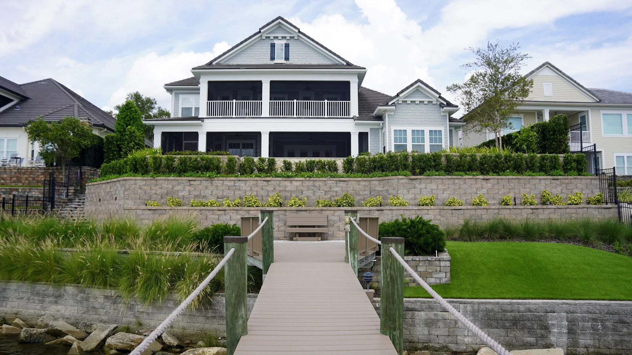 Large multi-story house with a front porch and balcony, situated on a terraced landscaped yard with hedges, bushes, and a small tree, with a pathway and railing leading down to a waterside with rocks.