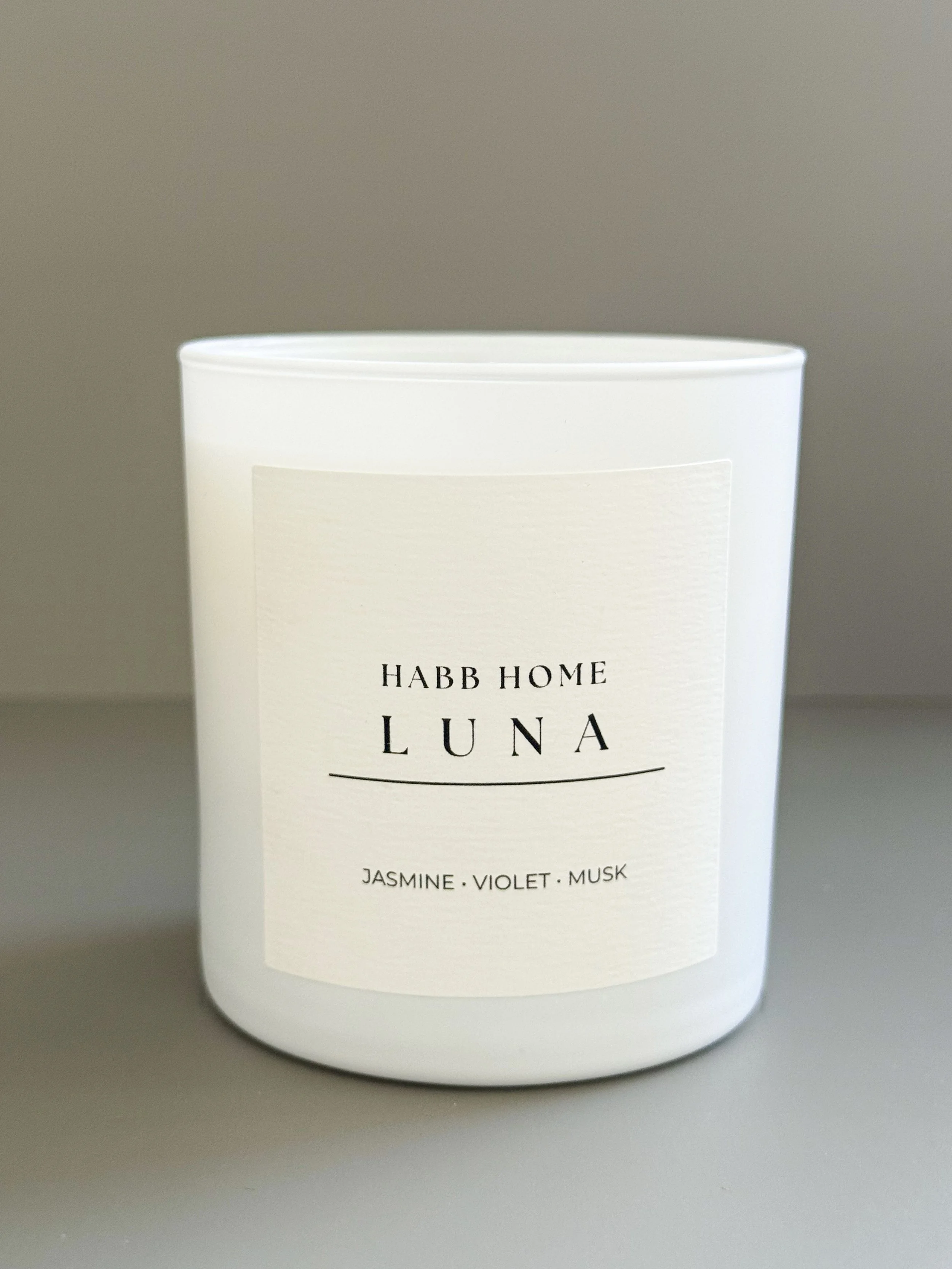 A white candle labeled "HABB HOME CANDLE" with scent notes jasmine, violet, and musk, surrounded by pink rose petals and dried lavender on a dark surface.