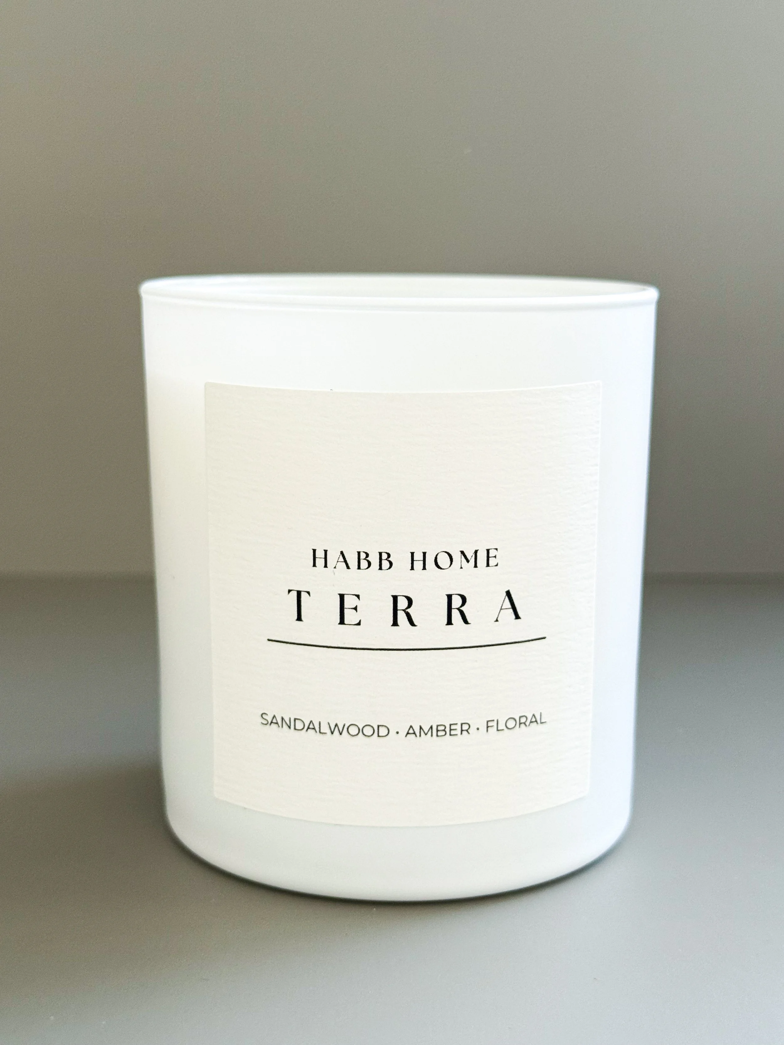 A white scented candle labeled 'Habb Home Candle' with notes of sandalwood, cardamom, and amber, placed on a peach-colored surface surrounded by dried flowers, cinnamon, and a brown textured box.