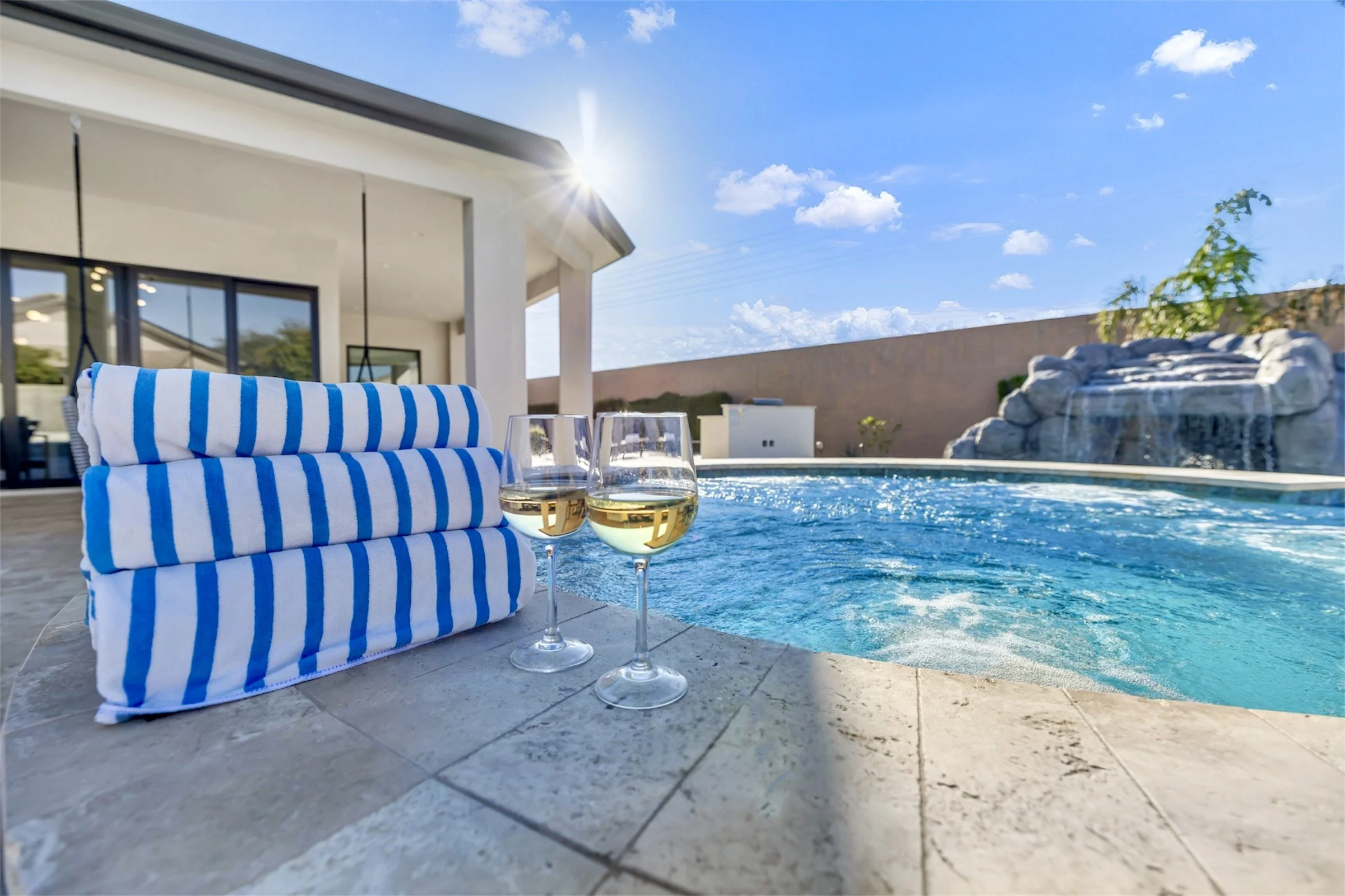 Close-up of striped blue pool towels and two wine glasses beside a sparkling backyard pool with sun flare and waterfall feature in Scottsdale, Arizona.