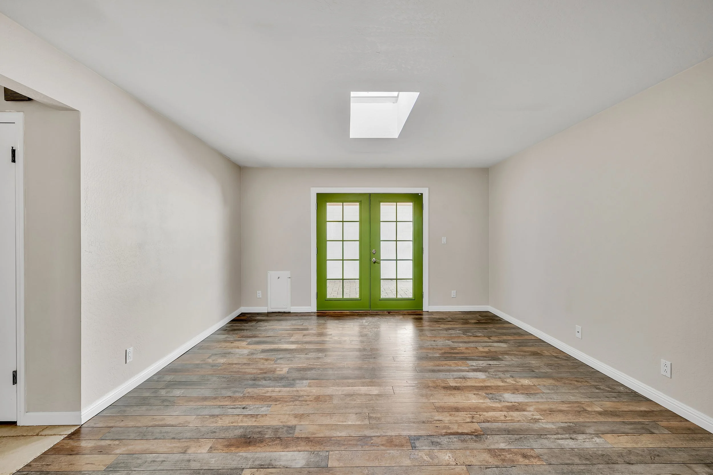 This bright empty room in Tempe, AZ features green French doors, skylight, and wood flooring, captured perfectly in real estate photography to showcase the space and natural light.