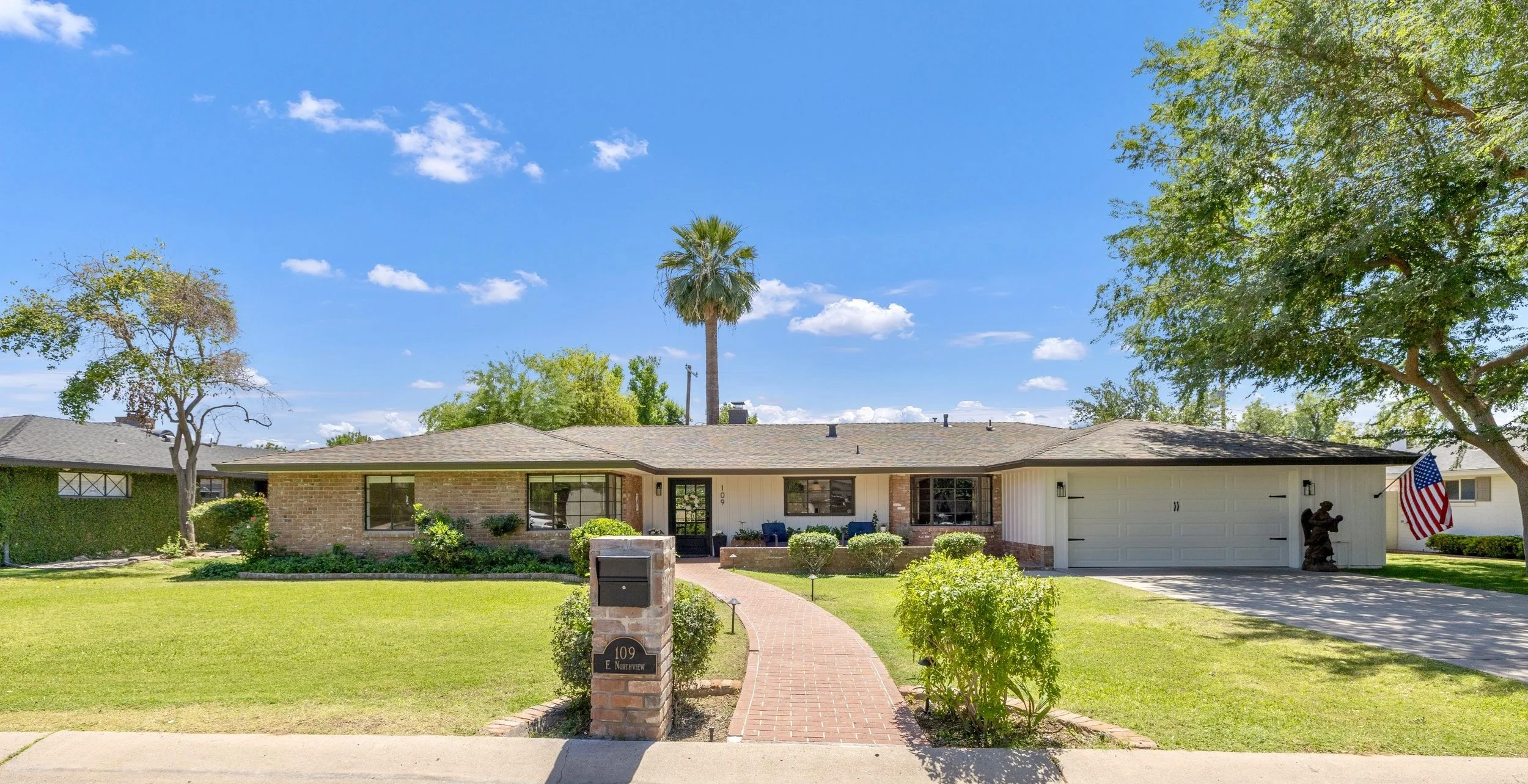 Real estate photography showcasing a charming single-story home on E Northview Ave in Phoenix, AZ, featuring a spacious front yard, brick walkway, and attached garage under a clear blue sky.