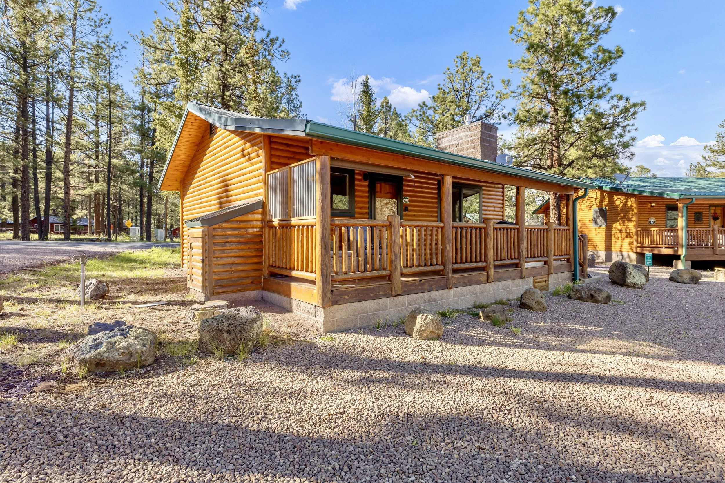 This charming log cabin with a wooden porch and gravel yard is showcased in professional real estate photography in Greer, AZ, surrounded by tall pine trees and natural beauty.