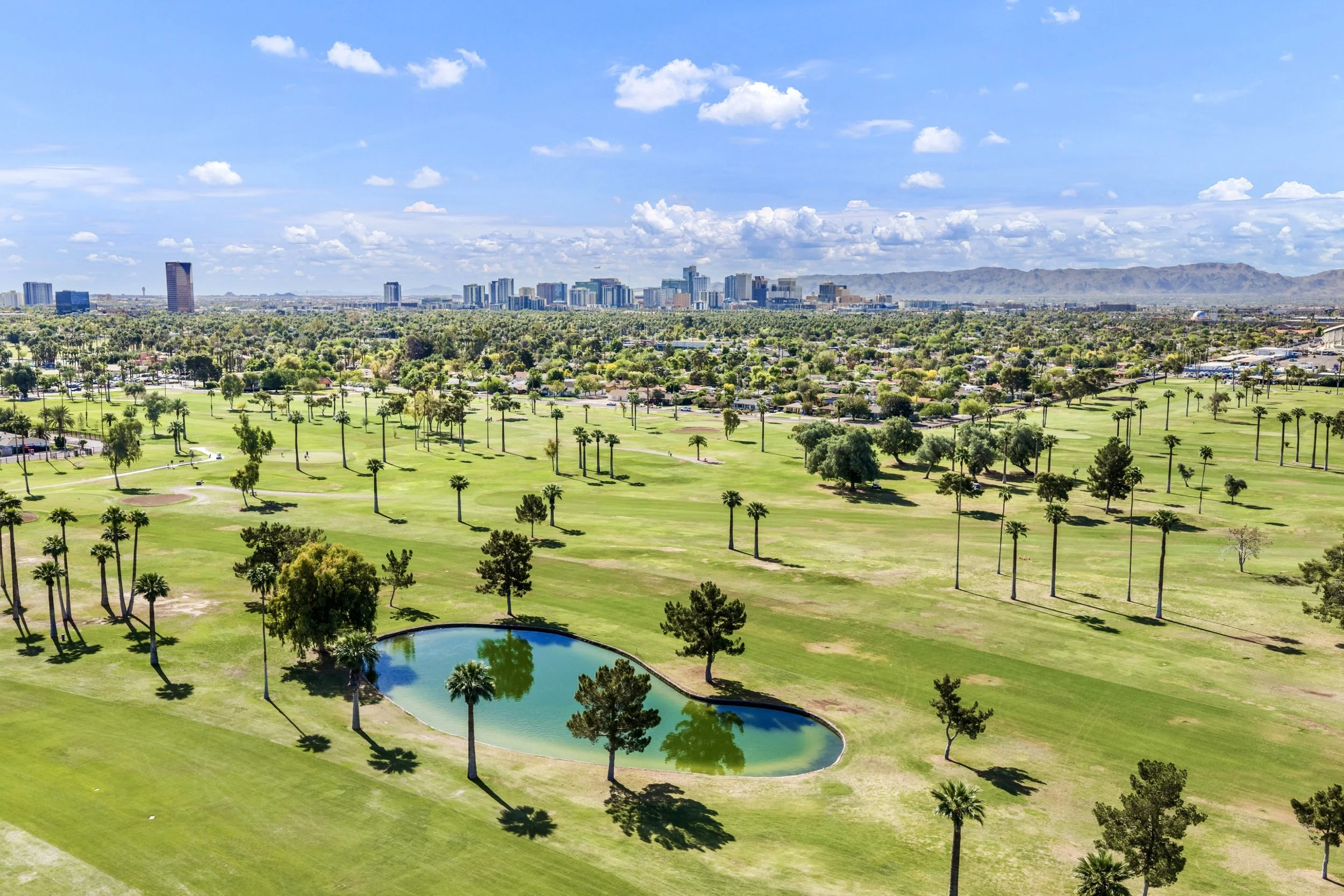 This aerial real estate photography captures a lush green golf course with a serene lake and palm trees in Phoenix, AZ, set against the stunning city skyline and mountain backdrop.