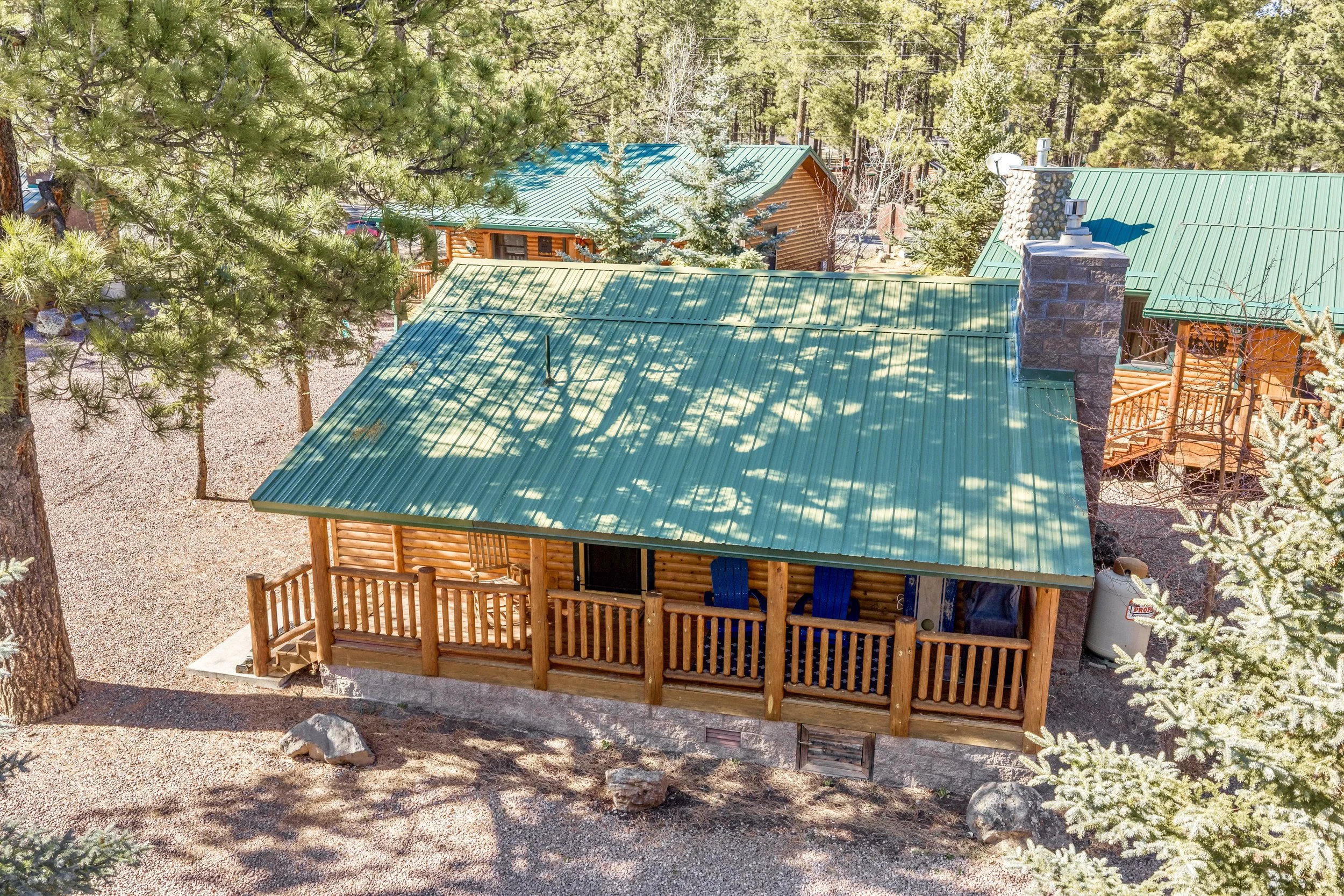 This aerial view showcases a charming log cabin with a green metal roof nestled among trees in Greer, AZ, highlighting the rustic appeal through professional real estate photography.