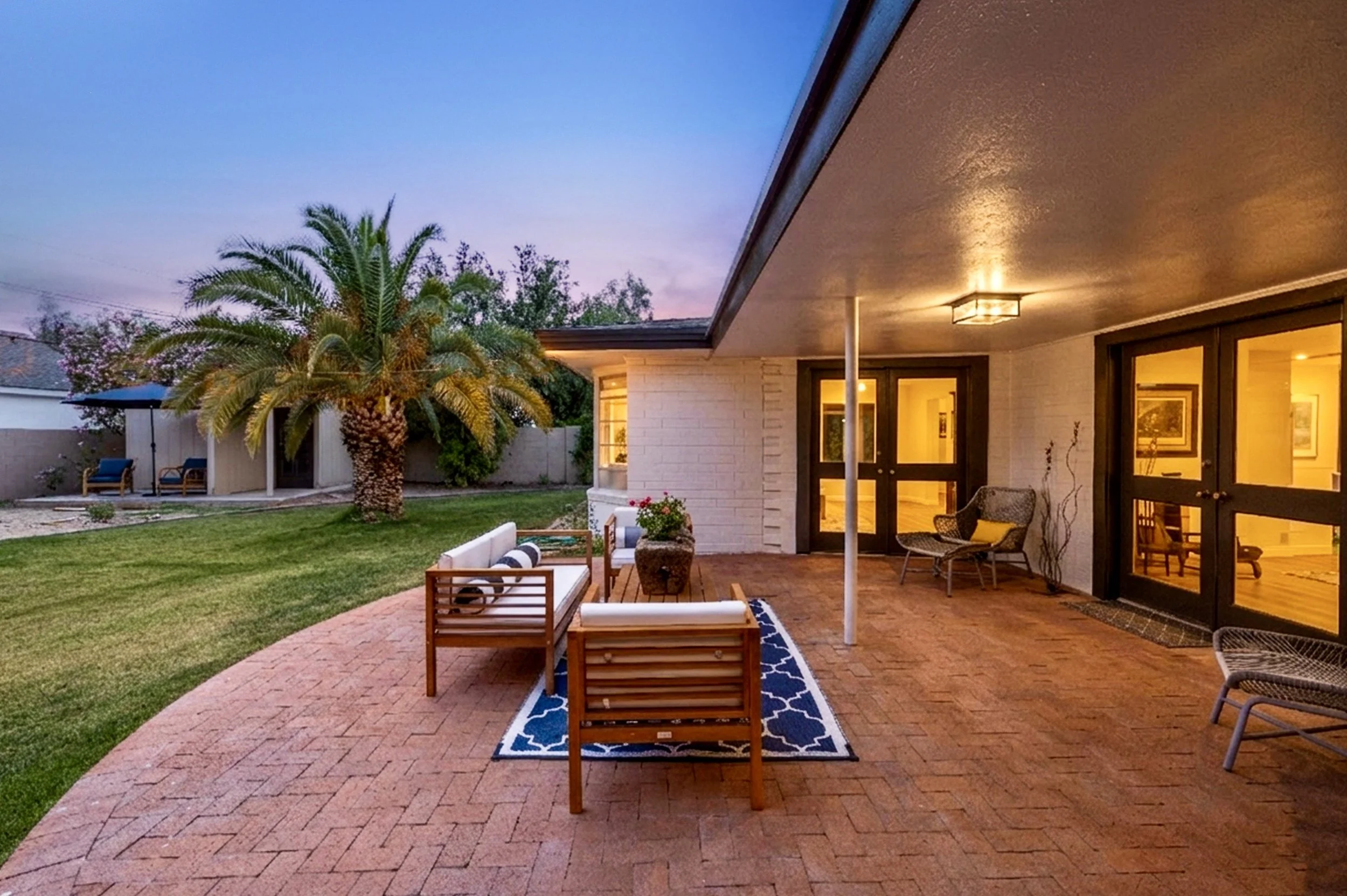 This beautiful backyard patio in Phoenix, AZ features cozy seating under a covered area next to lush greenery and a palm tree, captured through expert real estate photography at twilight.