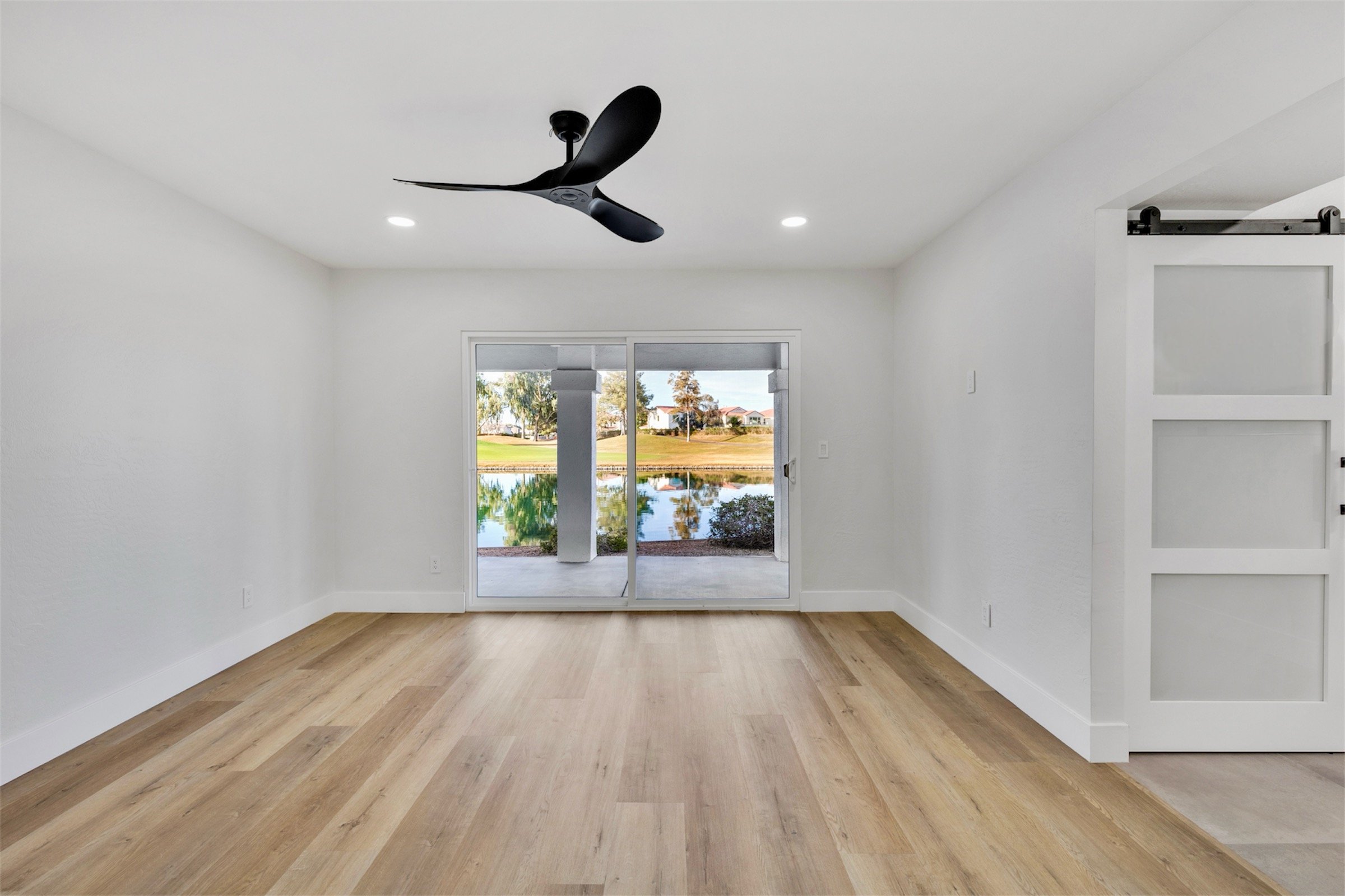 This empty living room in Chandler, AZ features wood flooring, a modern ceiling fan, and sliding glass doors showcasing a scenic water view captured with professional real estate photography.