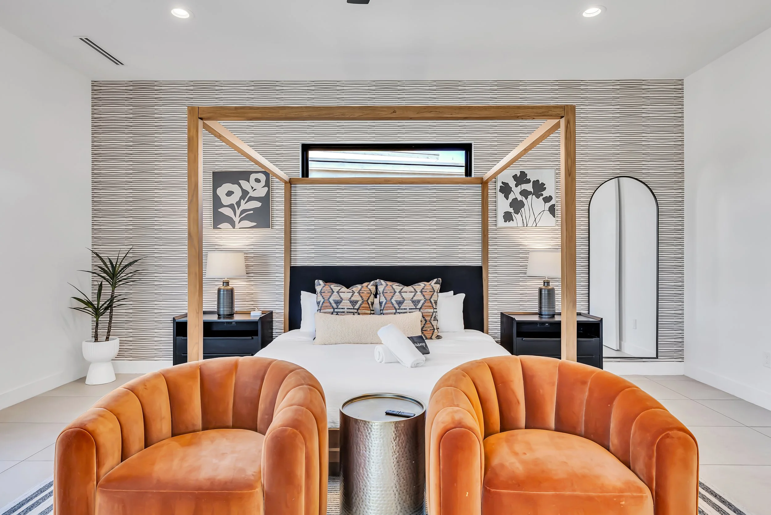 Front-facing view of the primary bedroom’s canopy bed with matching nightstands, botanical wall art, and two orange chairs in the foreground in Scottsdale, Arizona.