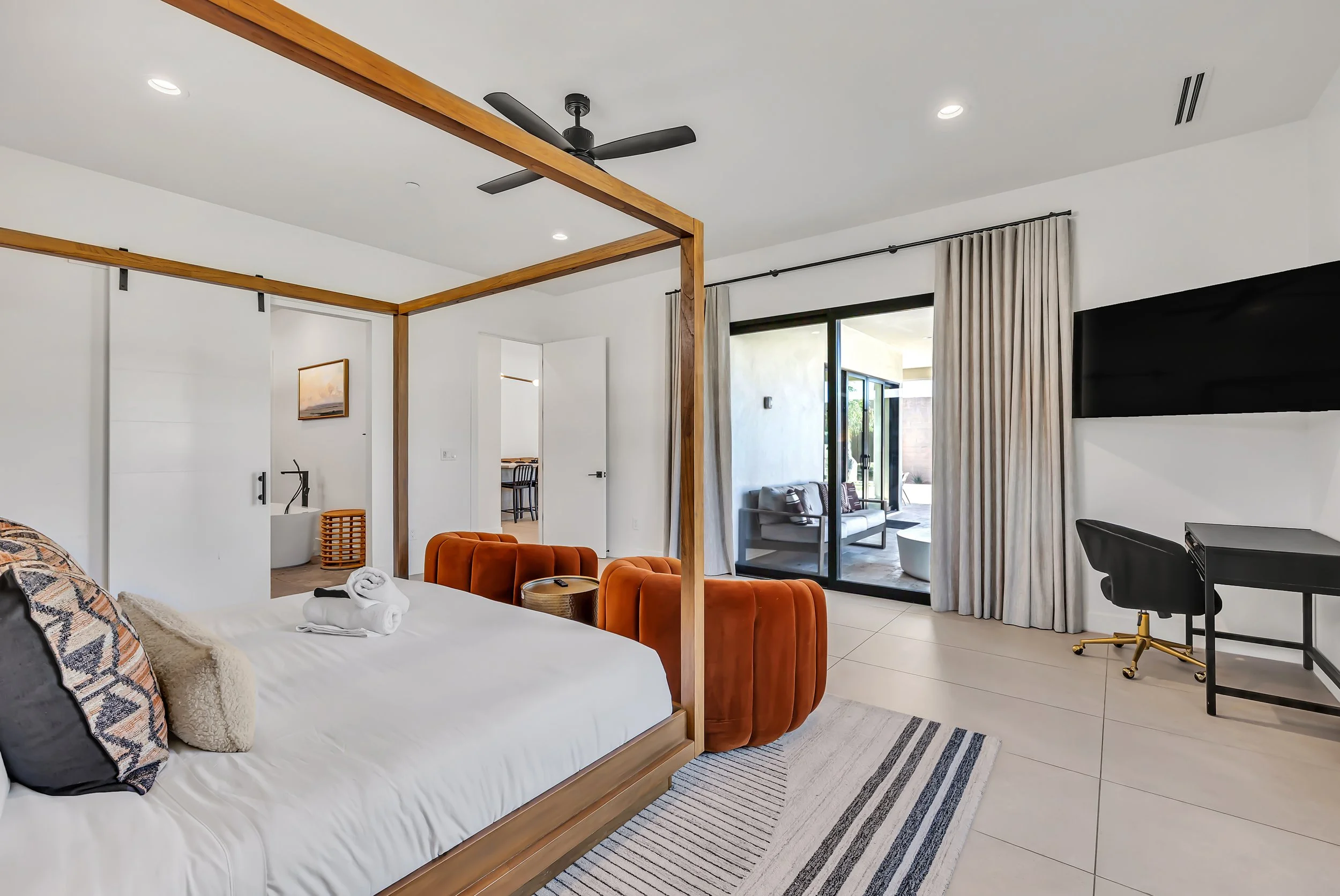 Primary bedroom angle showing canopy bed, orange chairs, sliding door to patio, and a small desk with a TV on the wall in Scottsdale, Arizona.