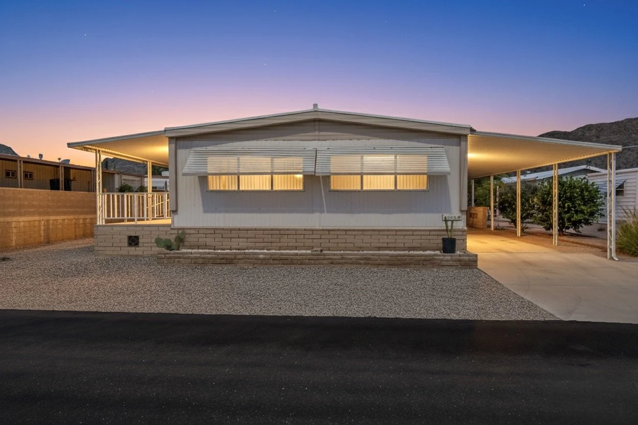This twilight exterior photo showcases a charming mobile home in Phoenix, AZ, illuminated warmly with covered parking and desert landscaping, perfect for real estate photography listings.