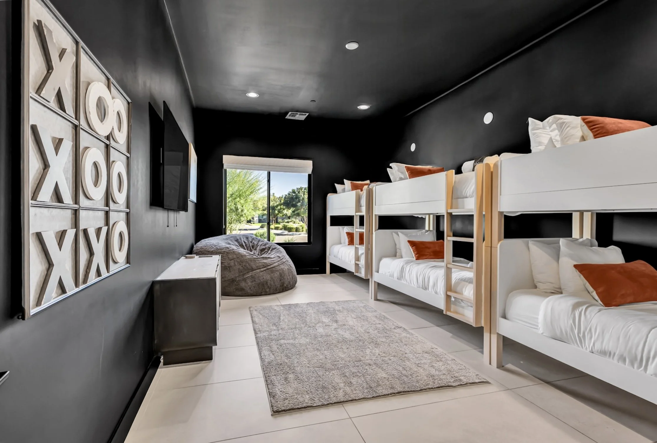 Stylish bunk room featuring four built-in beds, black walls, a wall-mounted TV, and a large window with natural light in Scottsdale, Arizona.