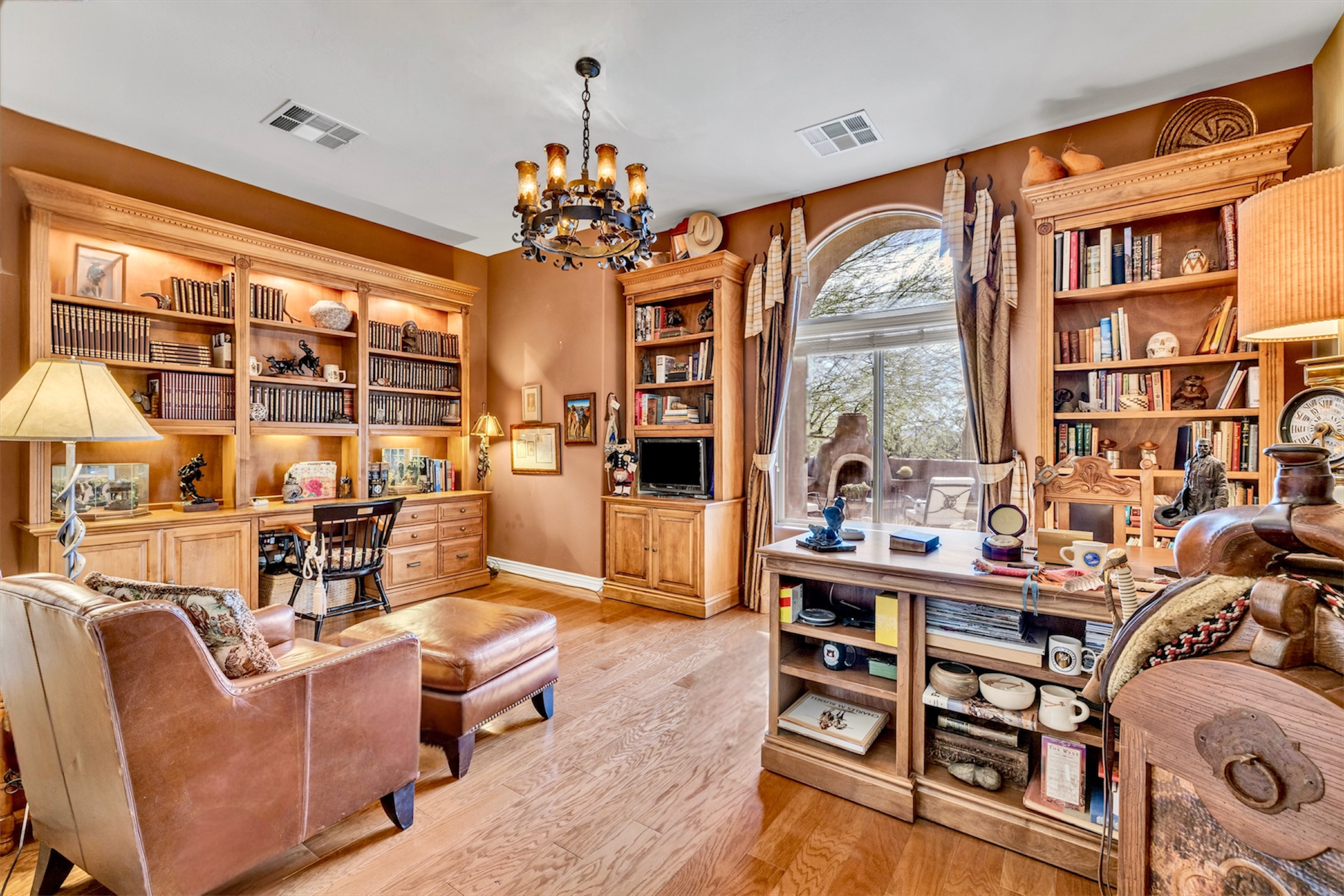 This Scottsdale, AZ home office features wood bookshelves, a leather chair, and a large arched window, perfect for a productive workspace captured in quality real estate photography.