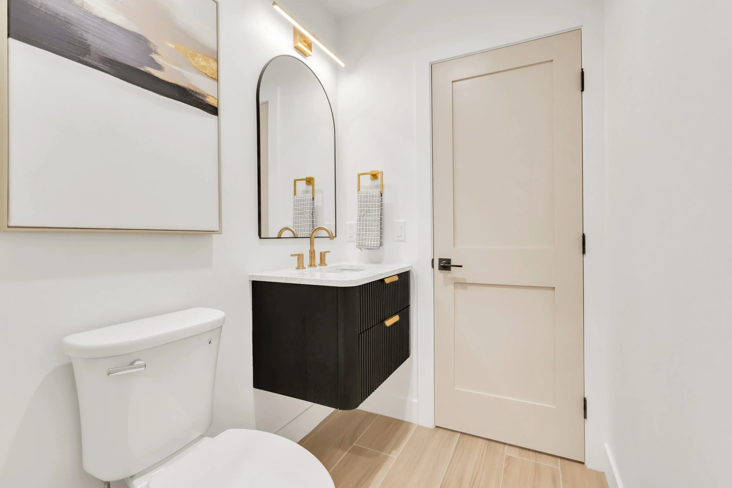 Stylish half bathroom in Phoenix, AZ with black floating vanity, gold fixtures, and arched mirror against white walls.