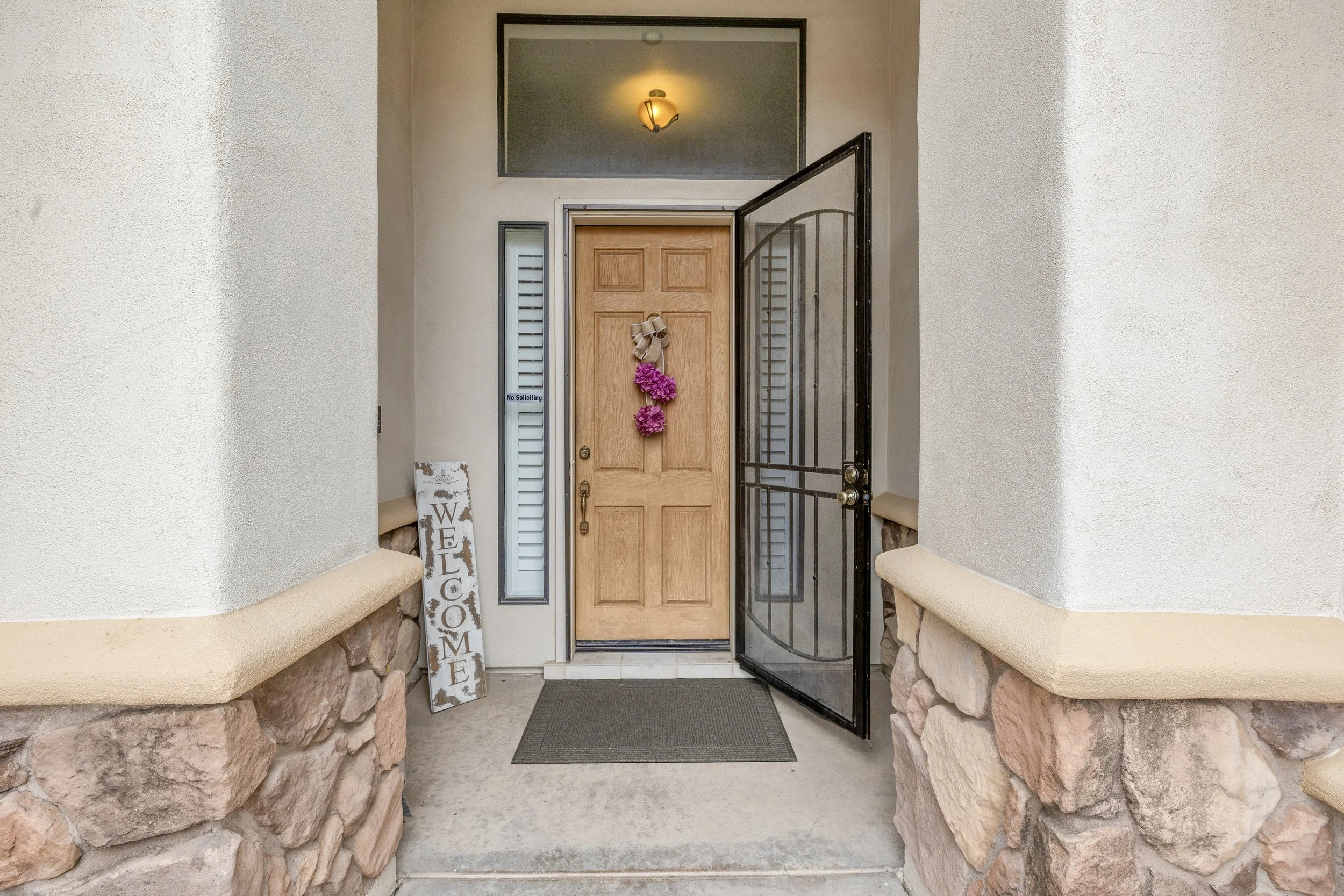 This Goodyear, AZ home features a charming wooden front door with decorative flowers and a welcome sign, highlighted through professional real estate photography to showcase warm curb appeal.
