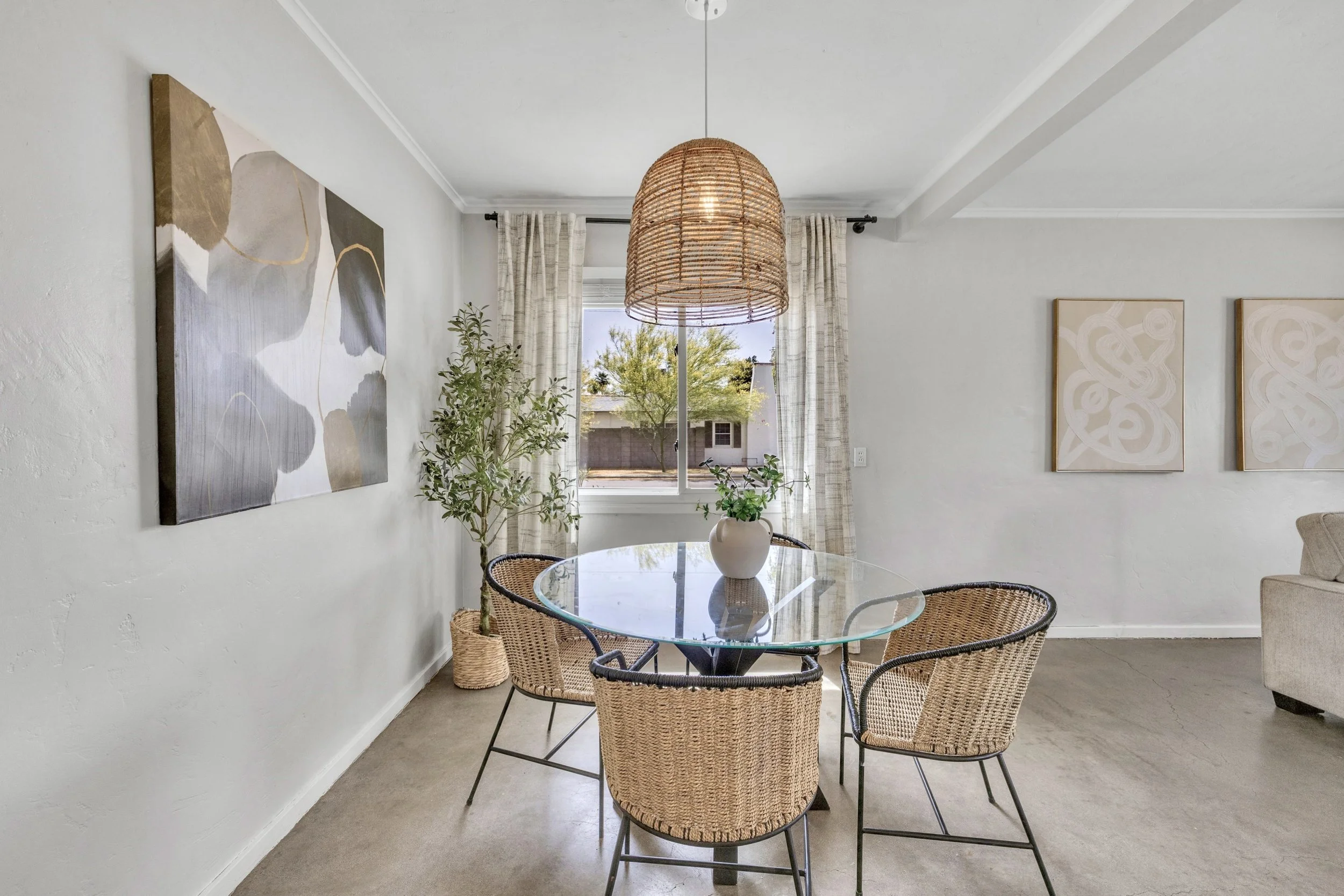 This modern dining room in Phoenix, AZ features natural light, a glass table, wicker chairs, and stylish decor, captured with professional real estate photography.