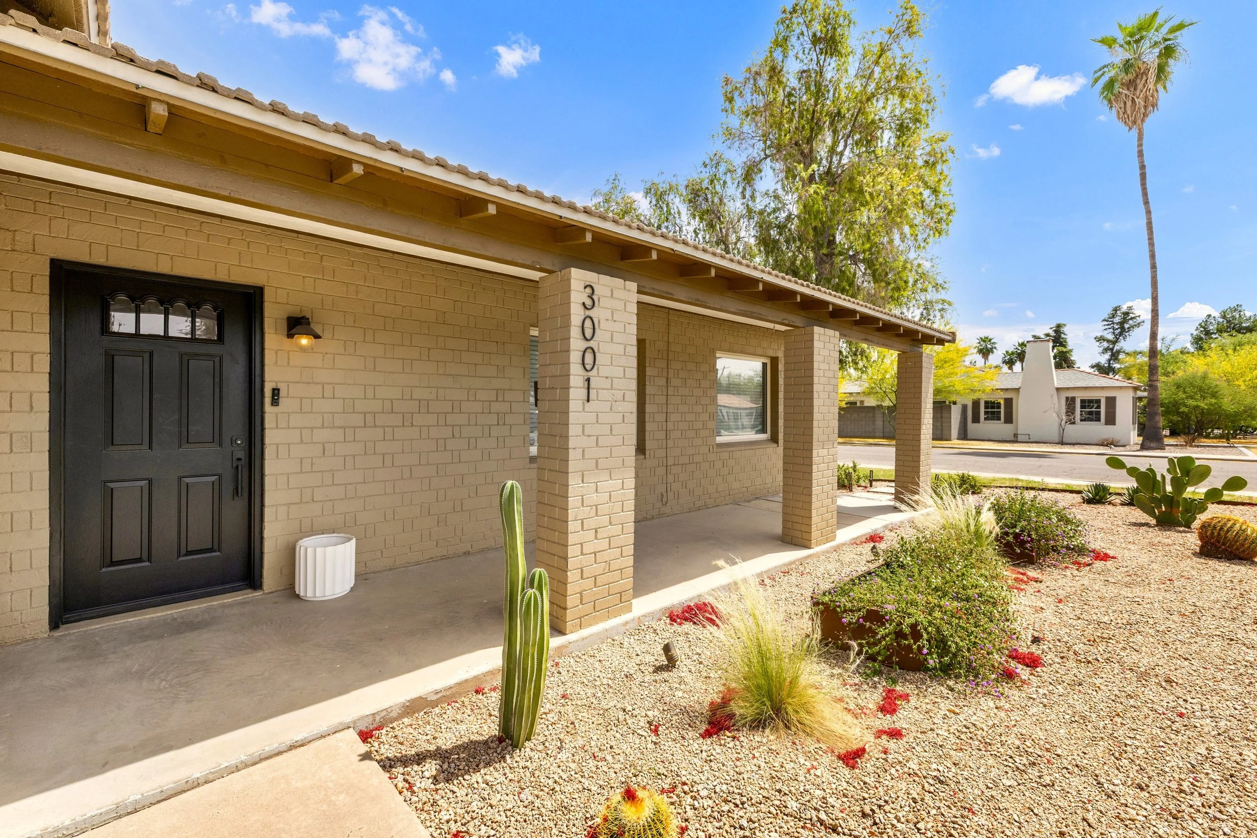 This real estate photography showcases a beige brick house with a covered porch and desert landscaping in Phoenix, AZ, highlighting its charming curb appeal under a clear blue sky.