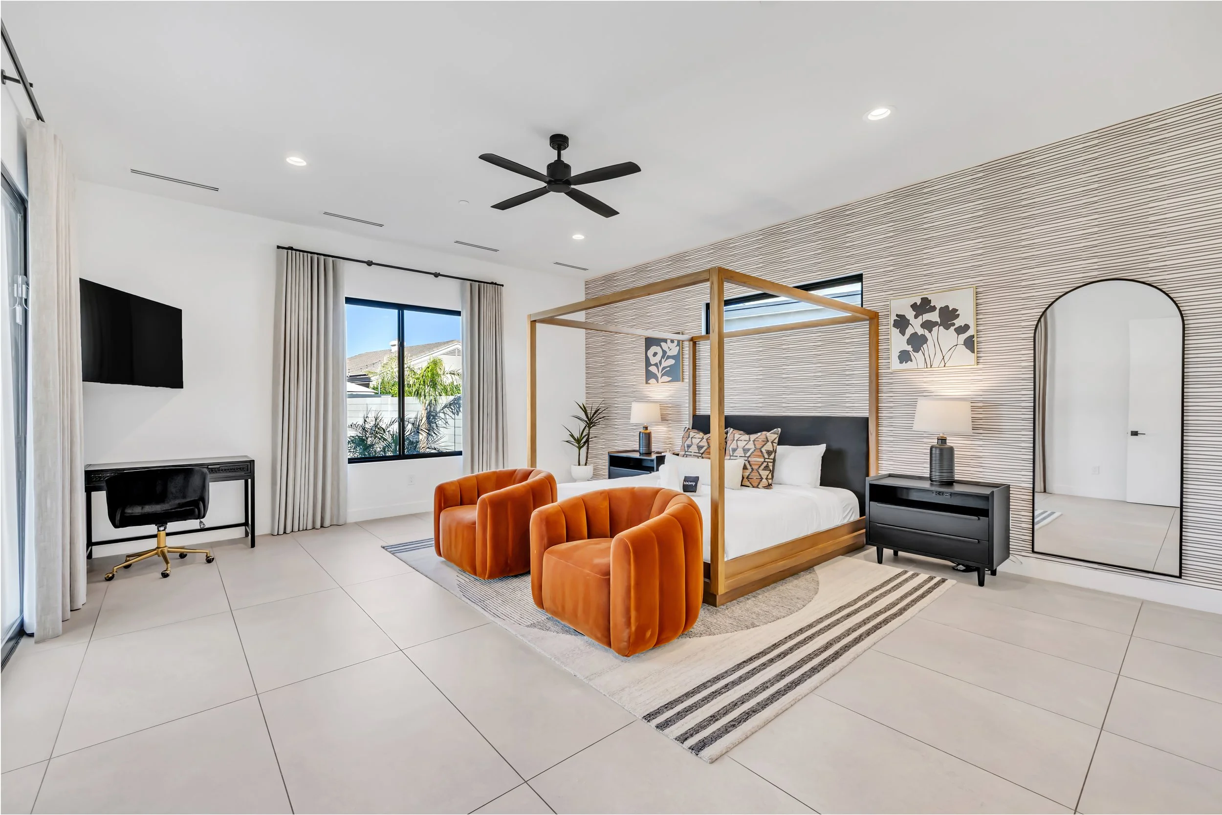 Large primary bedroom with a wooden canopy bed, modern wallpaper accent wall, two orange velvet chairs, and natural light from large windows in Scottsdale, Arizona.