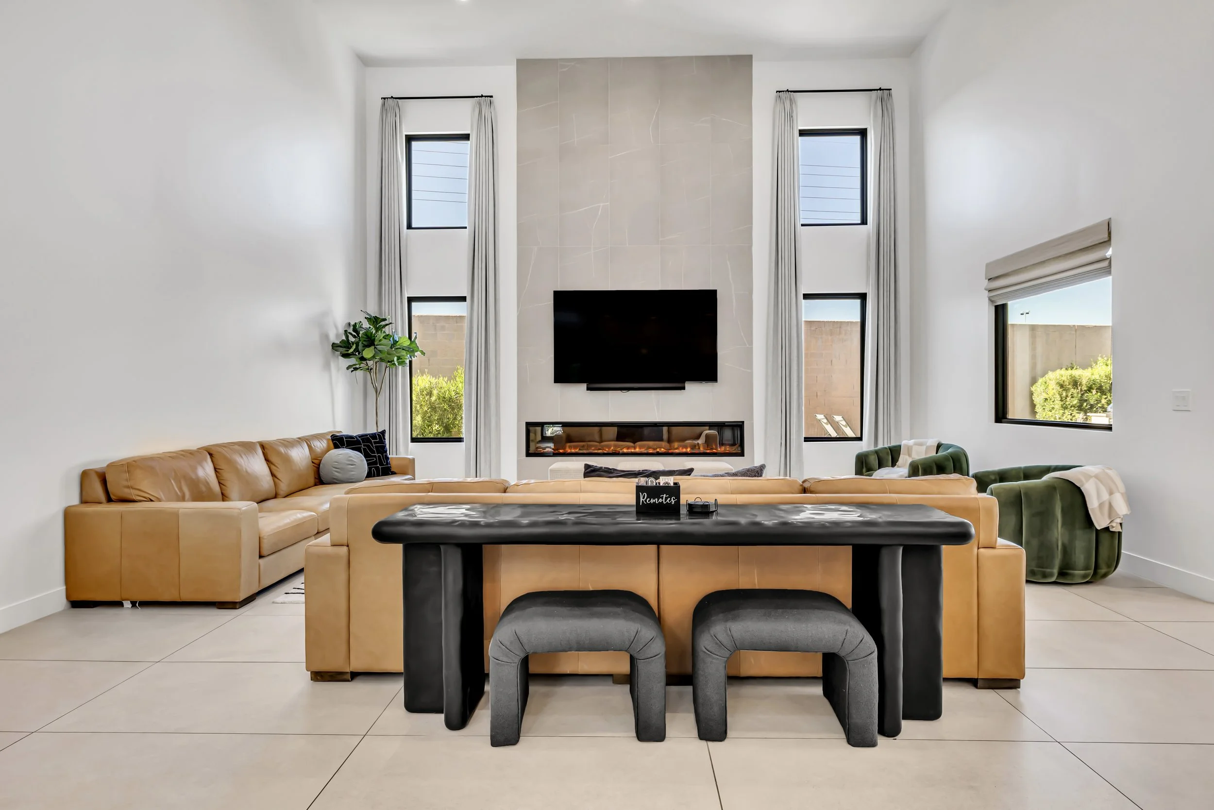 Wide shot of a living room with tan leather sofas, a fireplace with mounted TV, tall windows, and a central console table with stools in Scottsdale, Arizona.