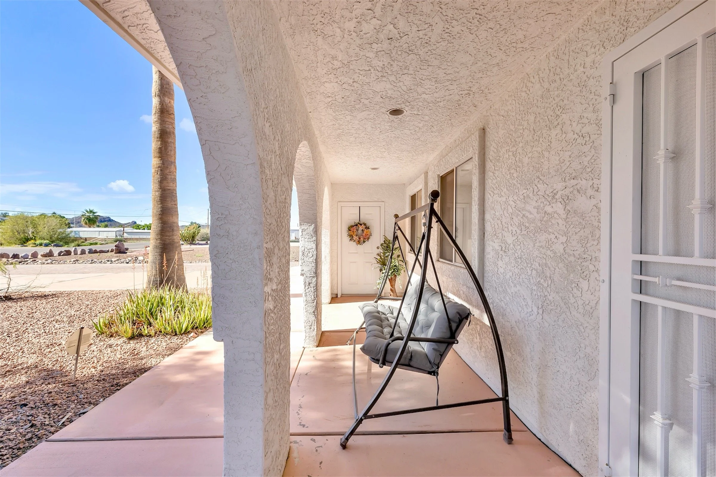 This real estate photography showcases a white stucco porch with arches and a cushioned swing in Queen Valley, AZ, highlighting inviting outdoor living spaces perfect for relaxation.