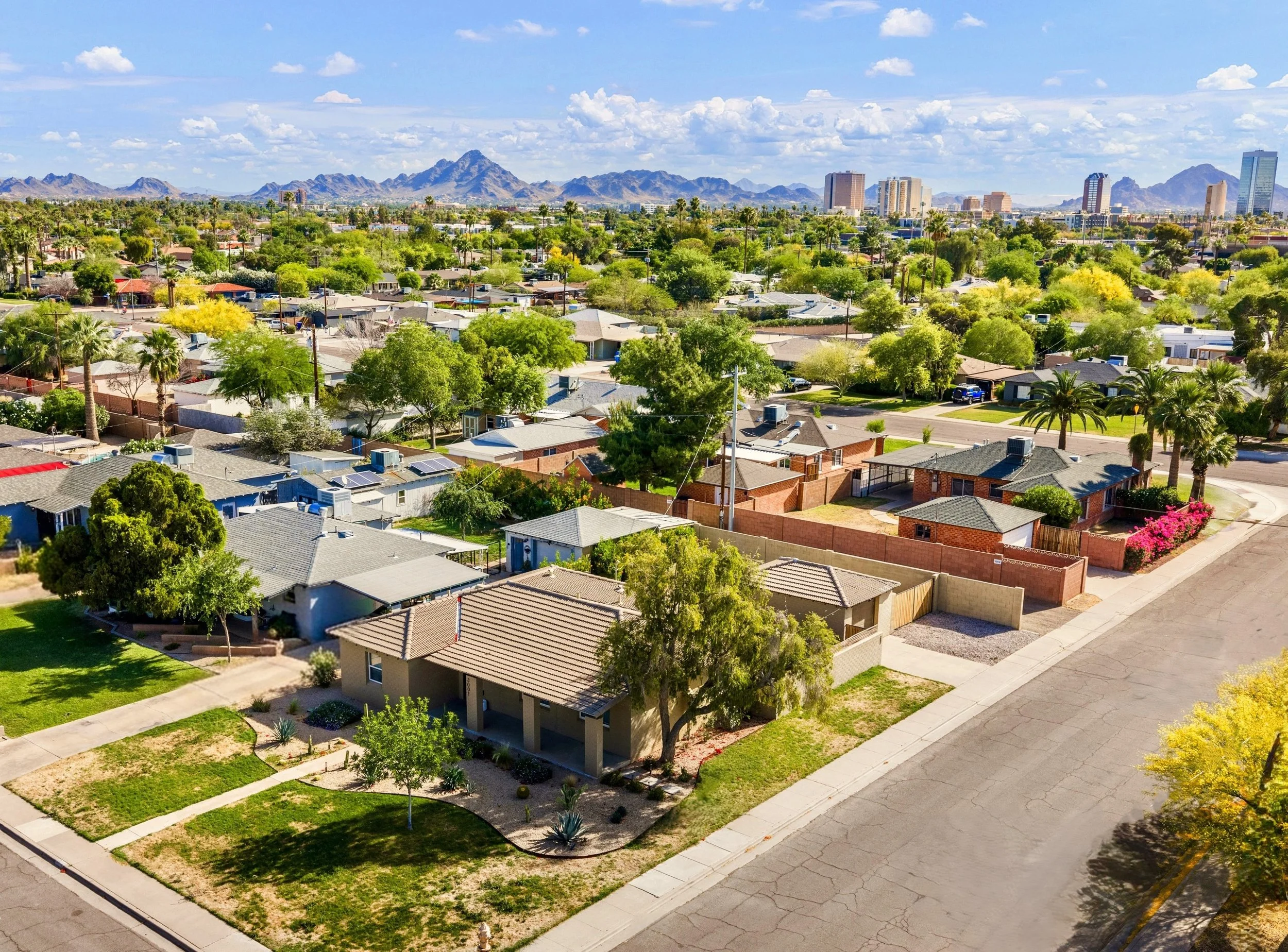 Aerial real estate photography capturing a Phoenix, AZ neighborhood with single-story homes, green trees, and mountains in the backdrop under a partly cloudy sky.