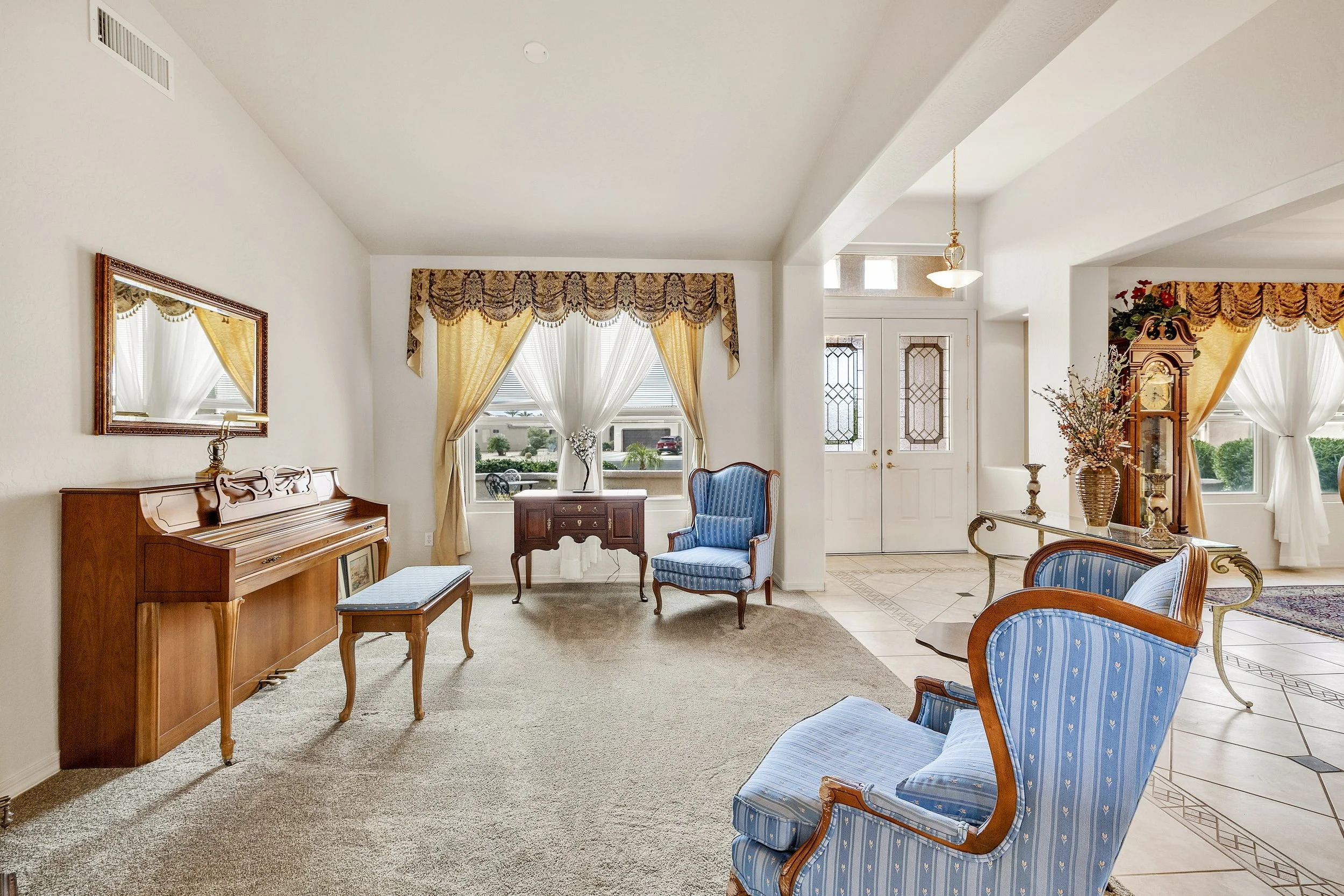 This elegant living room in Surprise, AZ, features a classic piano, blue armchairs, and bright windows, captured expertly with real estate photography to highlight its inviting charm.