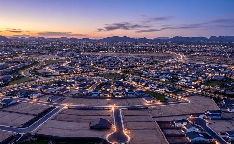 Aerial sunset view of an Arizona suburban neighborhood with city lights and mountain horizon.