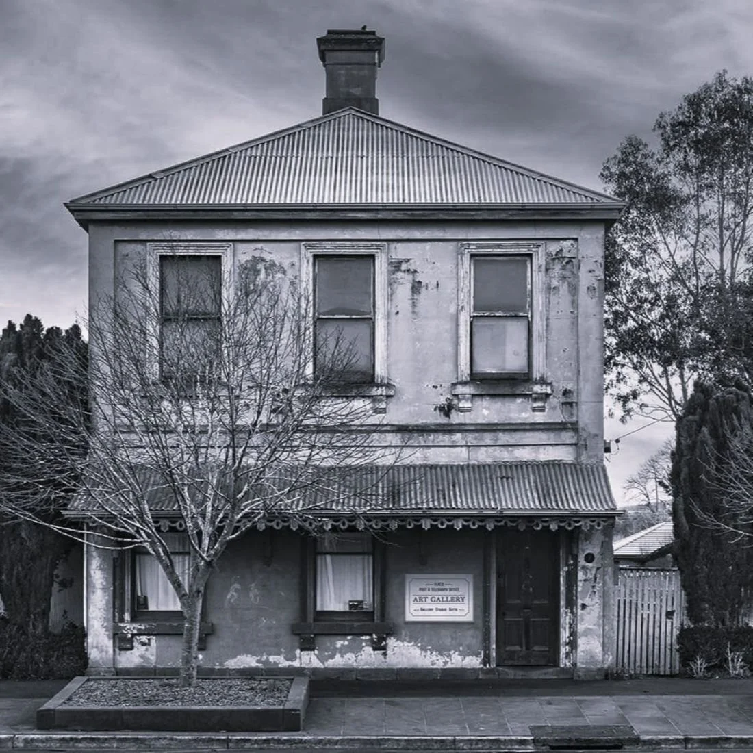 An old, weathered two-story building with a sign indicating it is an art gallery, with a leafless tree in front and a cloudy sky overhead.