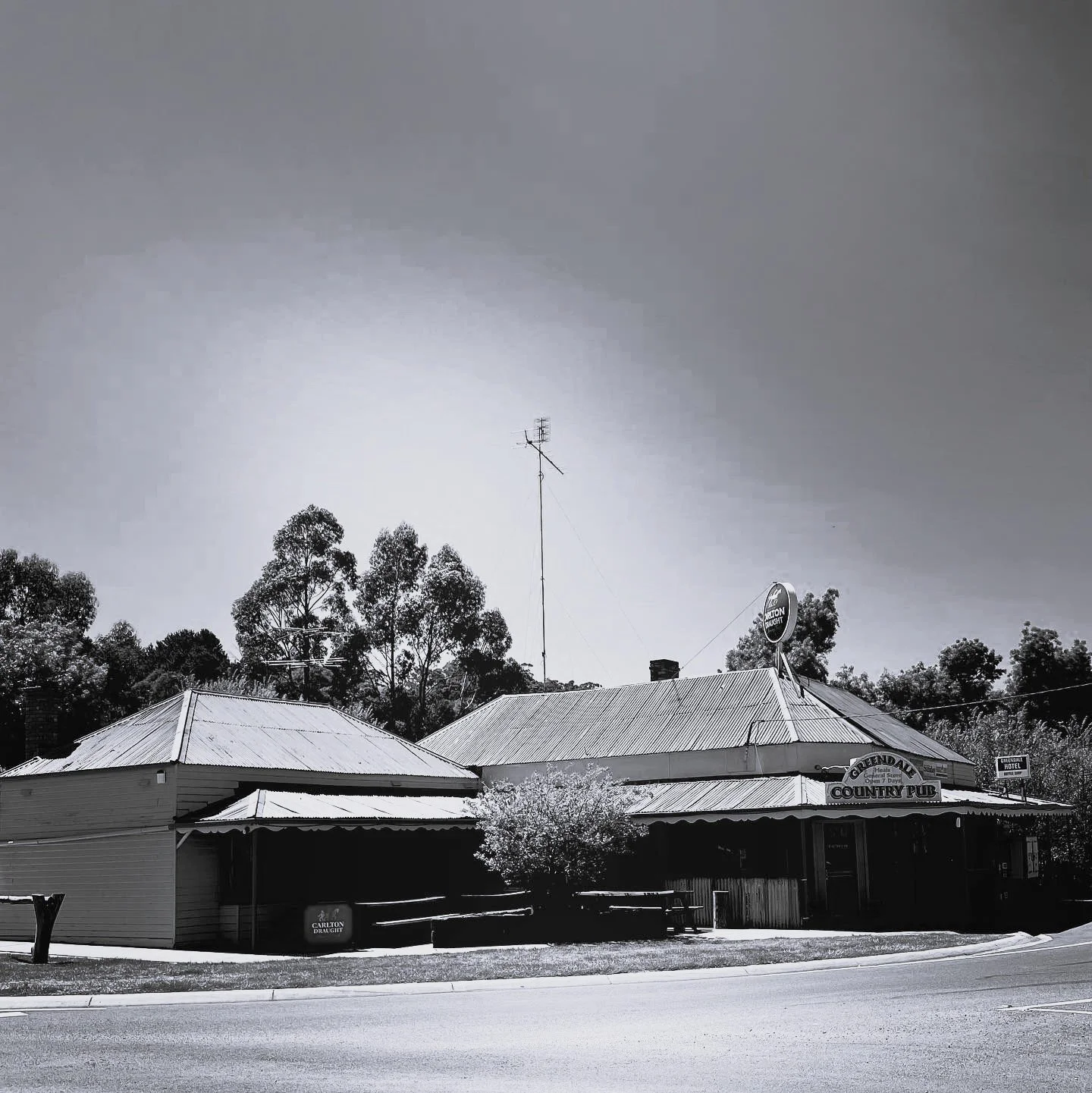 A black and white photo of a rustic country pub with a sign that reads 'Greendale Country Pub,' situated along a road with trees in the background.