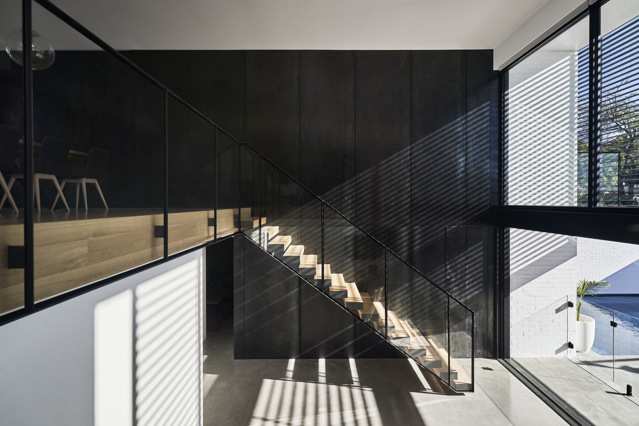 Modern interior space with staircase, glass railing, black wall, large windows with blinds, sunlight casting striped shadows, concrete flooring, white walls, and a potted plant outside.