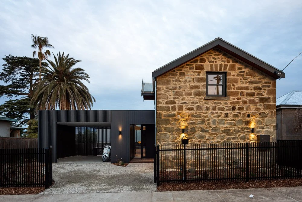 A house with a stone exterior and a black metal fence, modern black entrance structure, palm trees in the background, and outdoor lighting.
