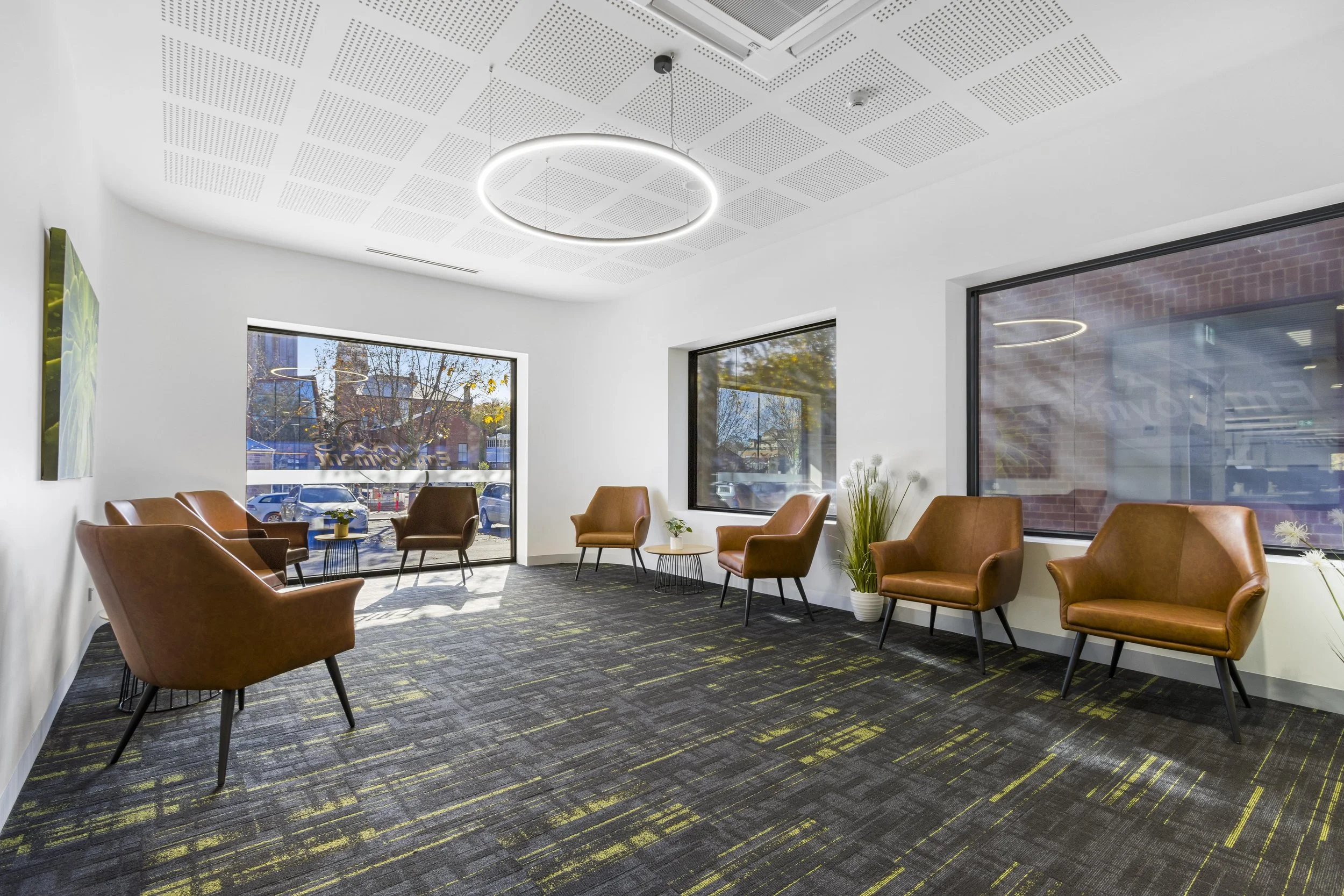 A modern waiting room with white walls and large windows, featuring six brown leather armchairs arranged around the room, a small round side table with a plant and books, a potted plant, and abstract wall art. Floor is carpeted with a dark, patterned rug, and a circular hanging light fixture illuminates the ceiling.
