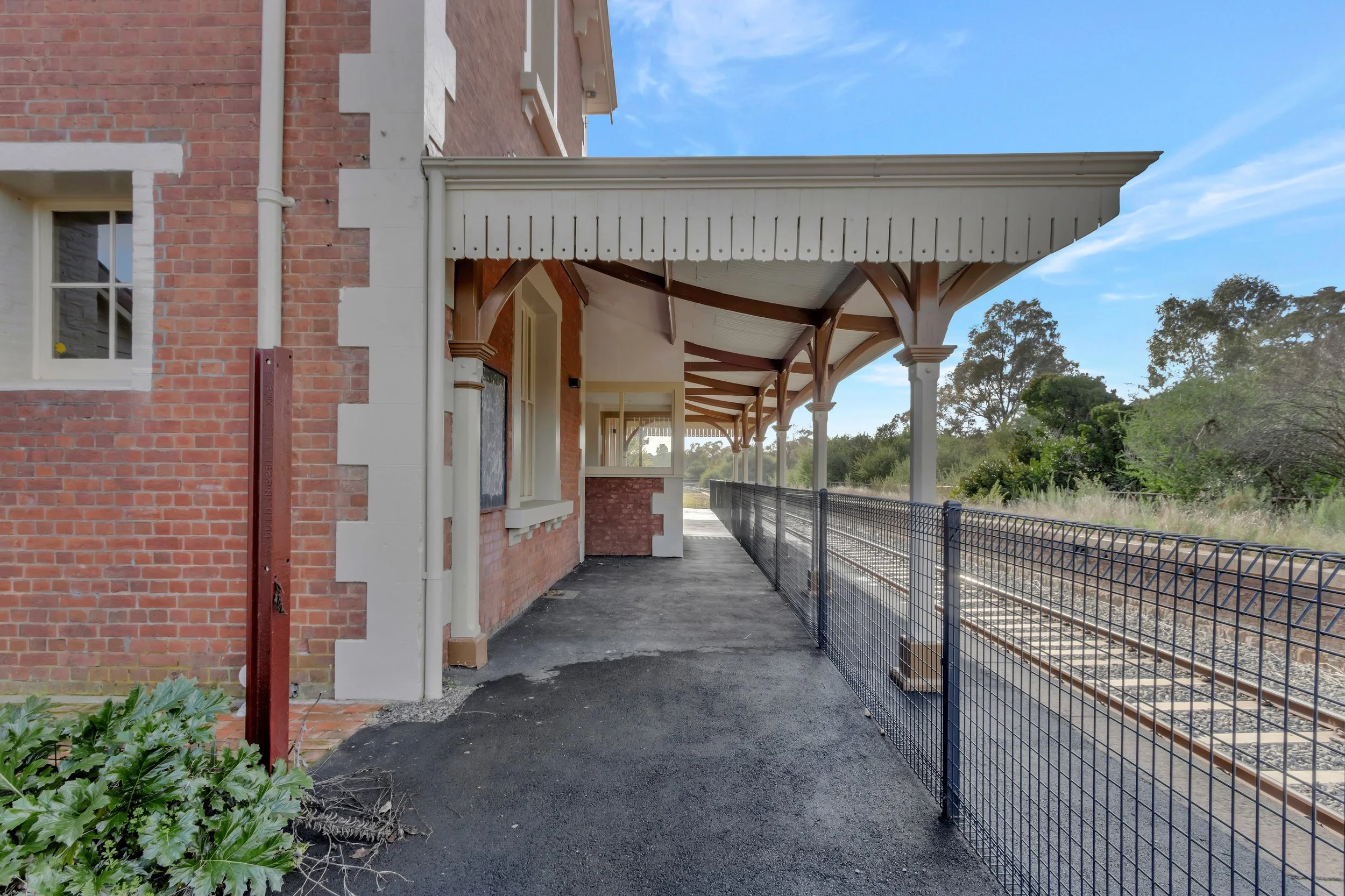 View of a vintage train station platform with a brick building, decorative wooden roof structure, and a metal safety fence along the train tracks, with trees visible in the background under a clear blue sky.