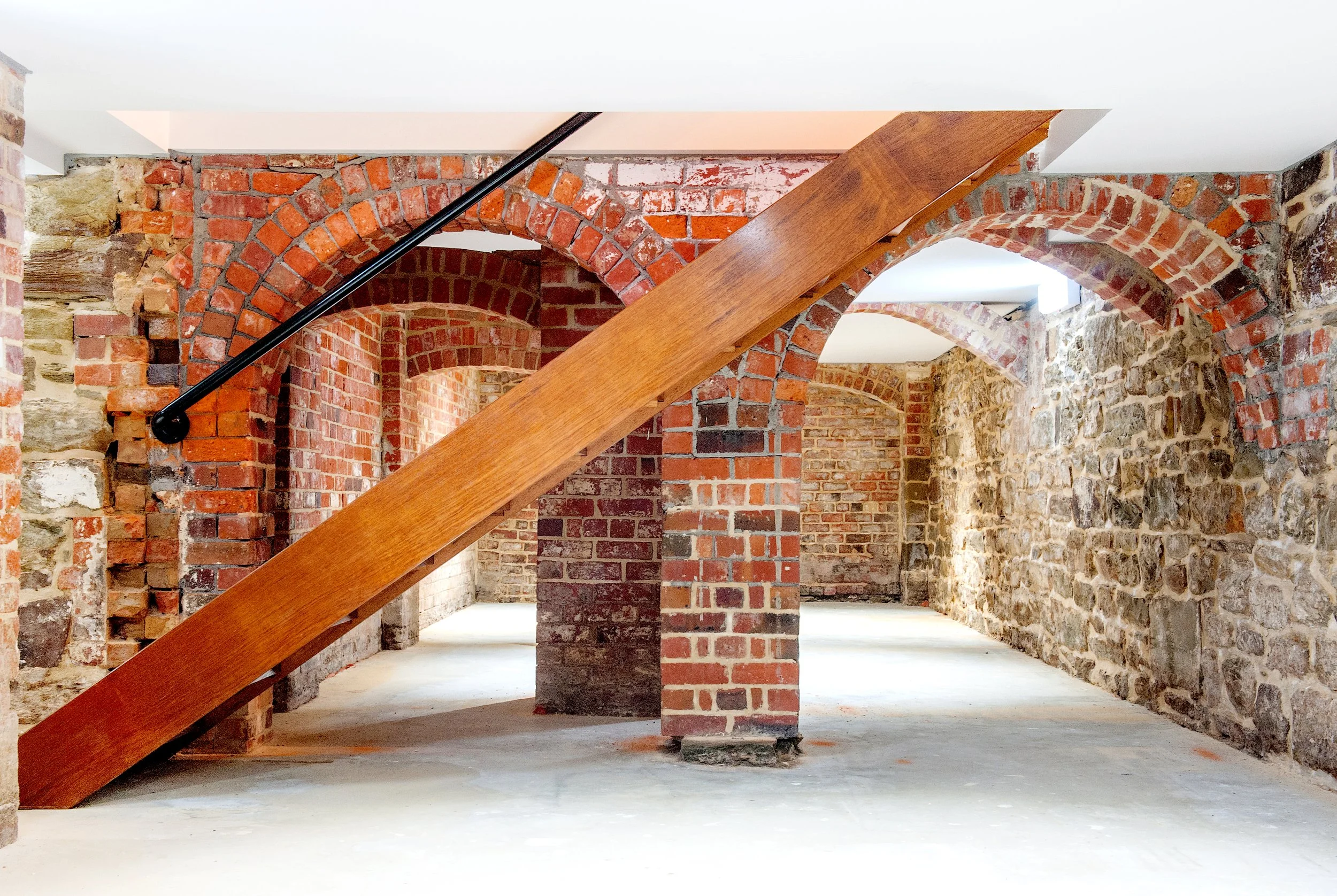 Interior of a basement or cellar with brick and stone walls, exposed brick arches, and a wooden staircase with a black handrail.