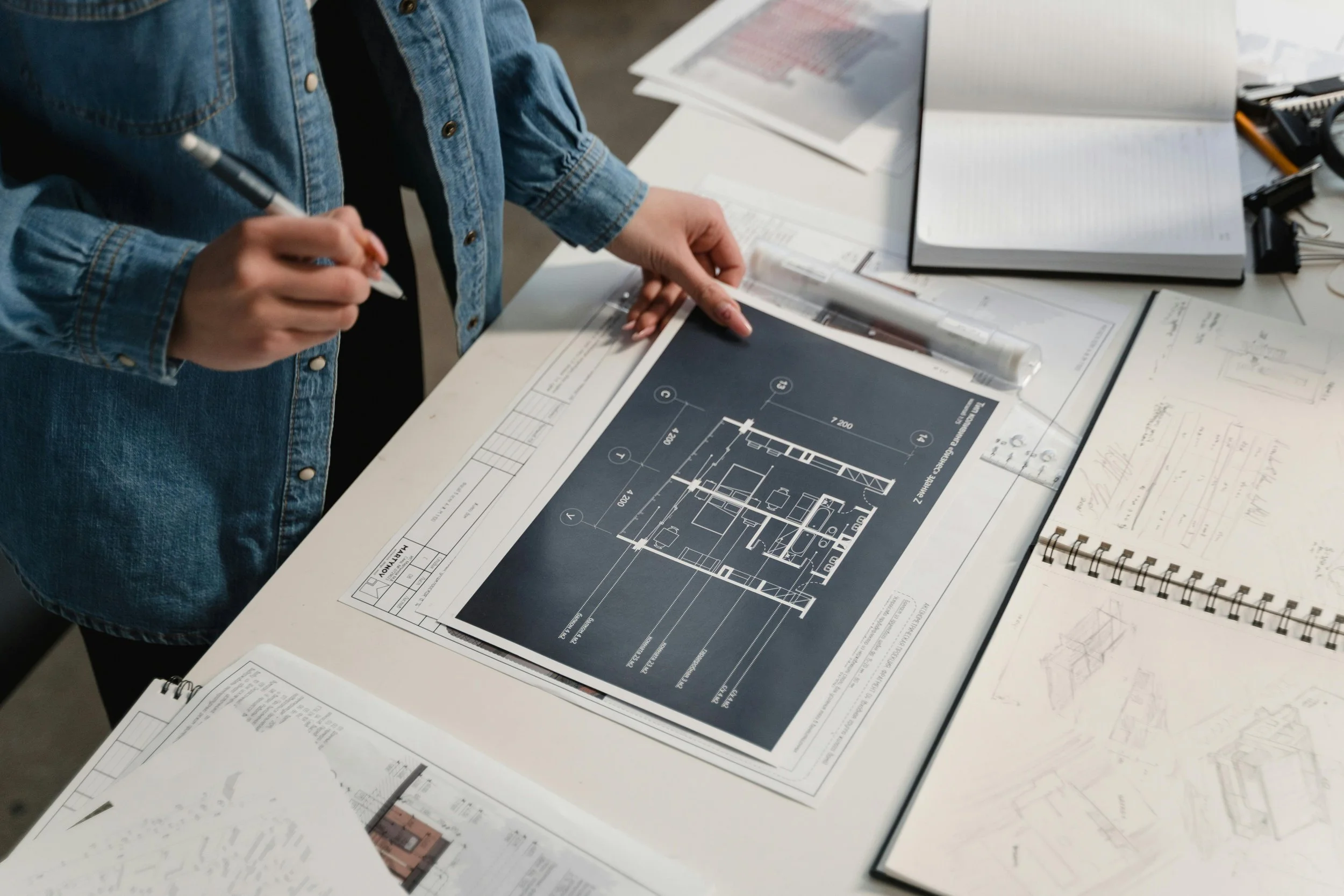 Person in denim jacket reviewing architectural plans on a table, surrounded by notebooks and measuring tools.
