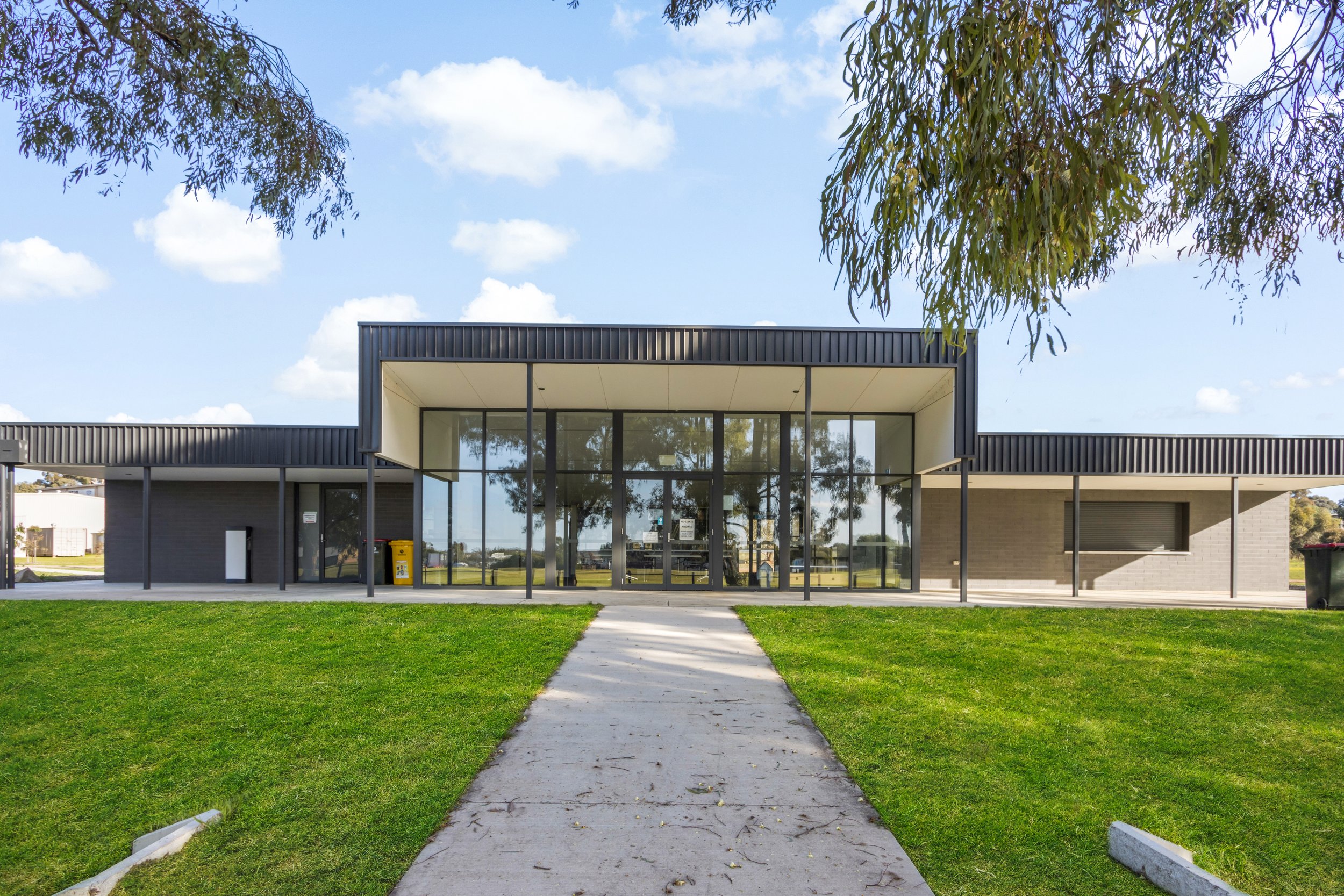 Modern building with large glass windows, surrounded by green grass and trees, under a partly cloudy sky.