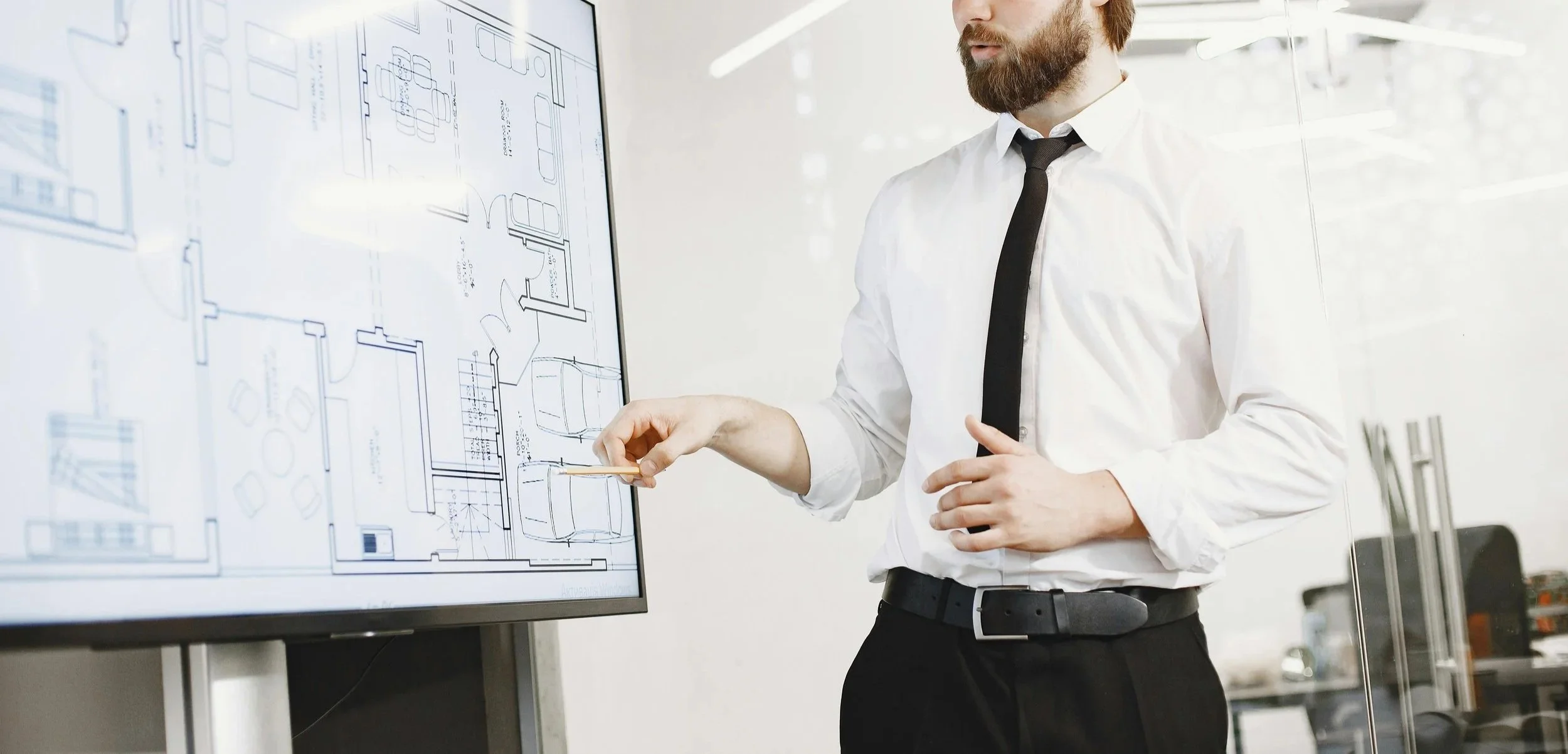 Man in white shirt and black tie presenting a blueprint on a large screen, discussing architectural or design plans.
