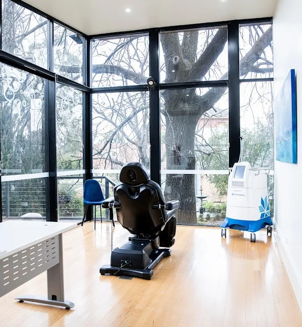 Medical room with a large window view of trees, a chiropractic or medical chair in the center, a blue chair beside it, and a medical device on wheels in the corner.