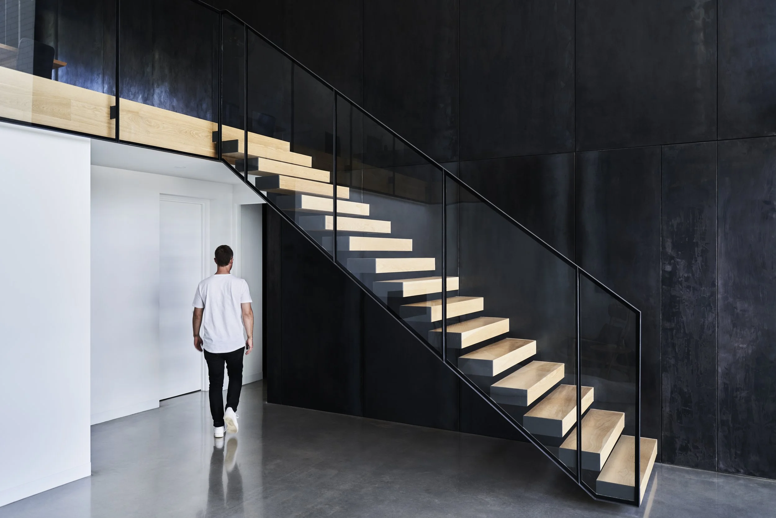 A man in a white t-shirt and black pants walking on a polished concrete floor toward a white door, with a modern staircase with wooden steps and black railings on a dark wall