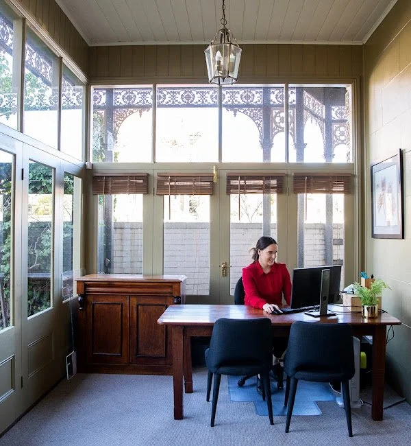 A woman sitting at a wooden desk in a sunlit room with large windows, working on a computer.