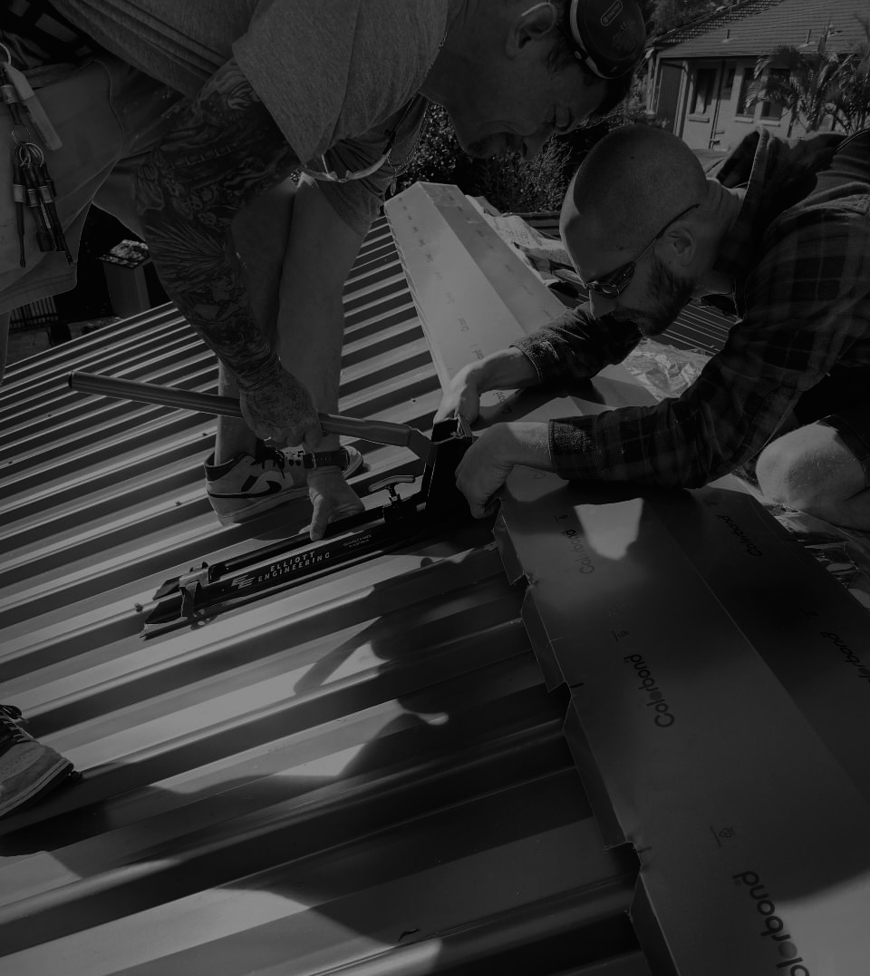 Two men working together on a metal roof, one is using a tool to secure the roofing panels while the other holds the panels in place, with a residential neighborhood visible in the background.